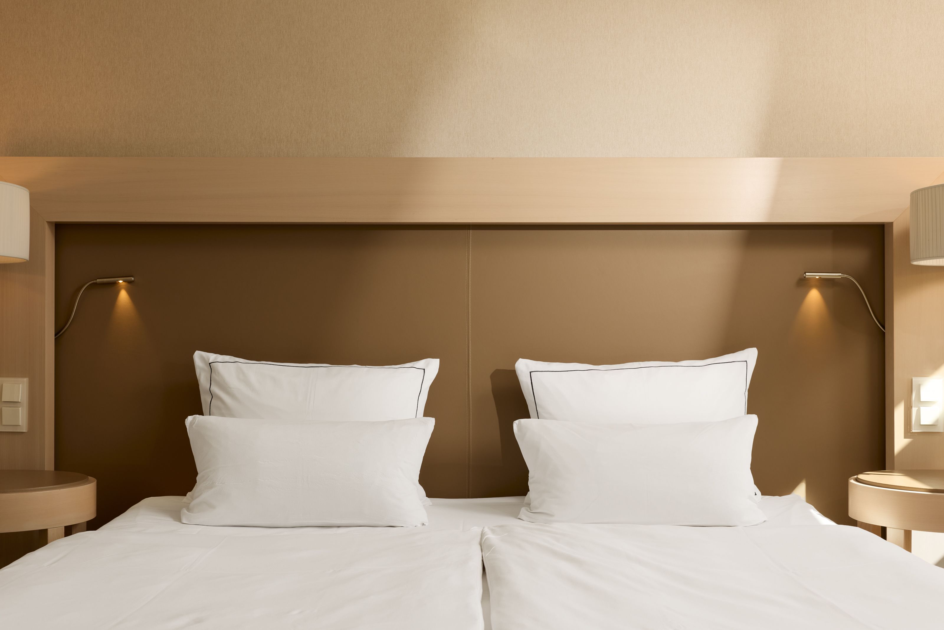 Zweibettzimmer mit zwei Einzelbetten, klarer, moderner Einrichtung in warmen Beige- und Brauntönen, jeweils mit Leselampe und Nachttisch. Ambiente vermittelt Ruhe und Komfort für erholsamen Schlaf.