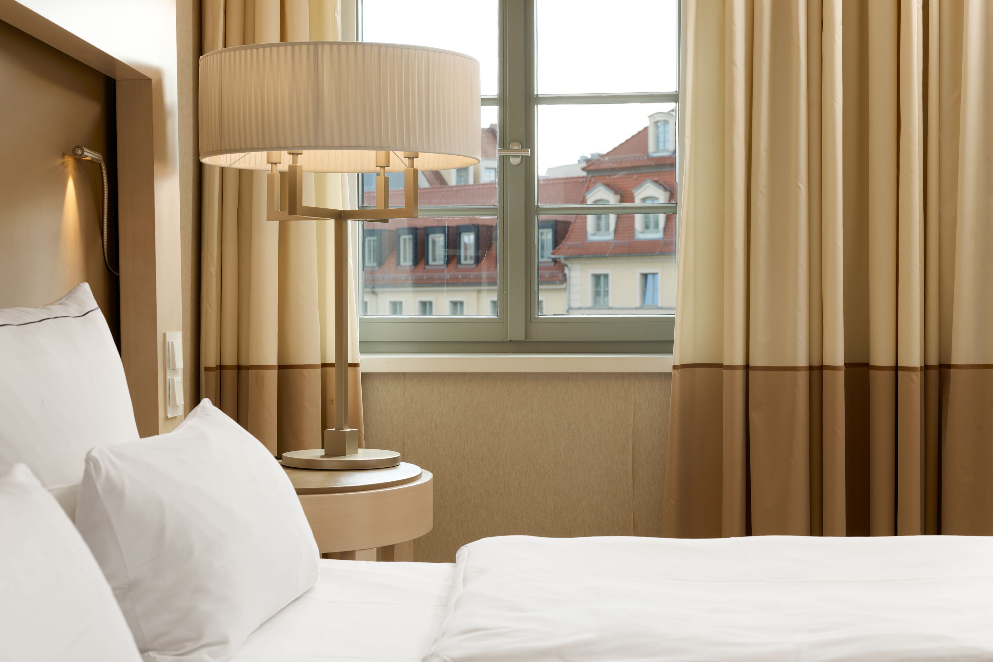 Hell eingerichtetes Hotelzimmer mit gemütlichem Bett, Nachttisch und Lampe, vor einem Fenster mit Blick auf historische Gebäude. Sanfte, warme Farbtöne schaffen eine einladende Atmosphäre.