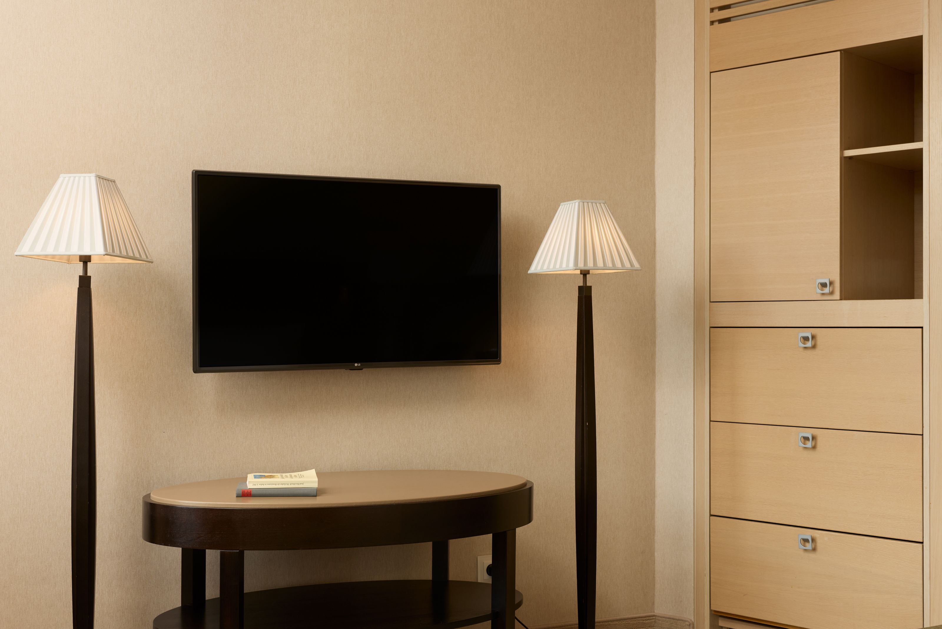 Modern eingerichteter Hotelzimmerbereich mit Flachbildfernseher an der Wand, zwei stilvollen Stehlampen und einem eleganten ovalen Beistelltisch, der eine einladende, ruhige Atmosphäre schafft.