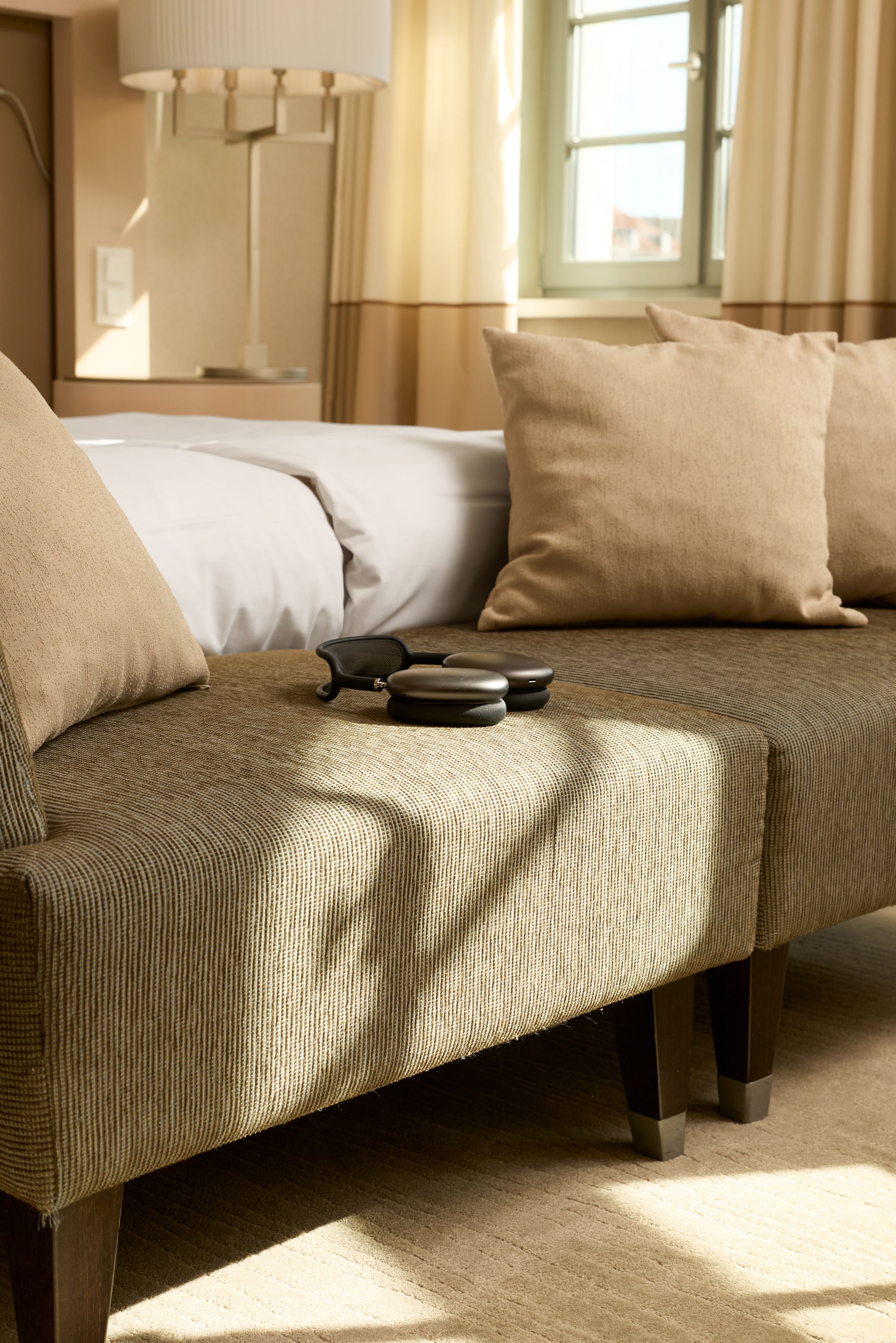 Helles Hotelzimmer mit bequemem, beigem Sofa und Kissen, auf dem eine Sonnenbrille und kleine Dosen liegen. Sanftes Tageslicht fällt durch die Fenster und schafft eine einladende, entspannte Atmosphäre.