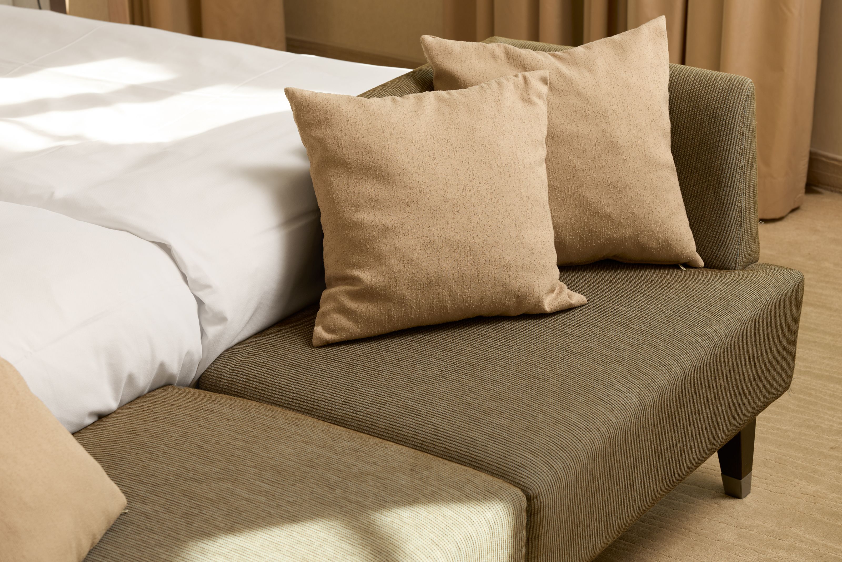Beiges Sofa mit zwei passenden Kissen neben einem weiß bezogenen Bett in einem hellen, komfortablen Hotelzimmer.
