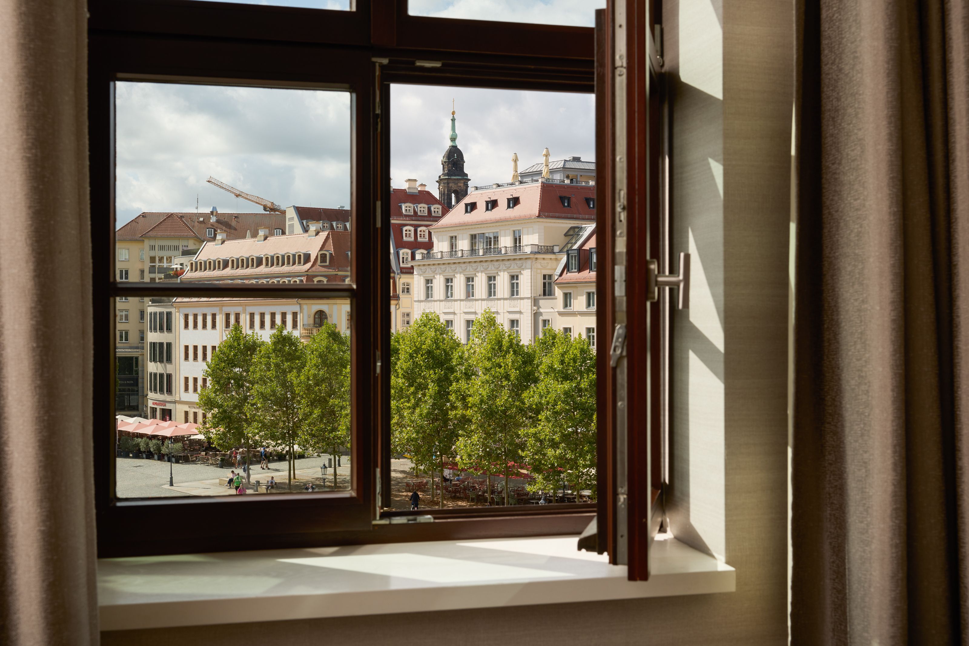 Blick aus offenem Hotelzimmerfenster auf historische Gebäude und grüne Bäume an einem städtischen Platz bei Tageslicht.