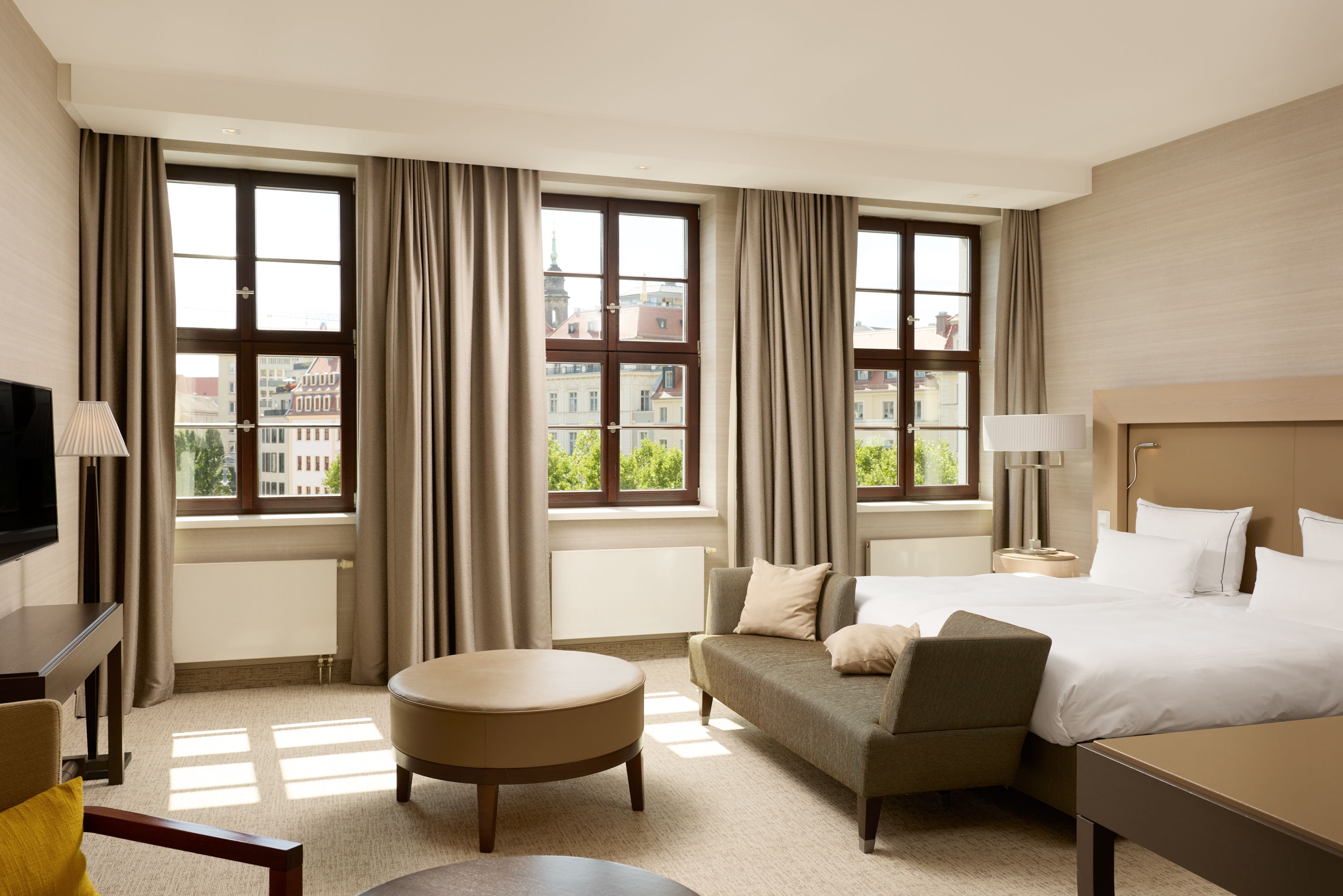 Helles, modernes Hotelzimmer mit großem Doppelbett, gemütlicher Sitzbank, runden Tischen und drei großen Fenstern mit Ausblick auf Stadtgebäude, Tageslicht durchflutet, warme, neutrale Farbtöne und komfortable Atmosphäre.
