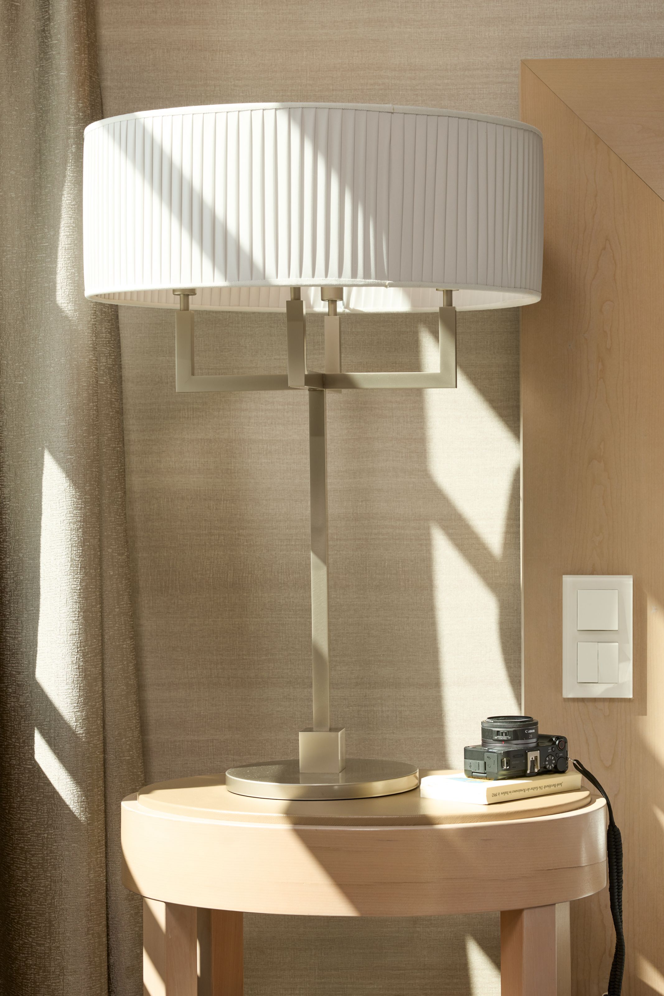 Moderne Tischlampe mit schirmförmigem weißem Lampenschirm auf rundem Holztisch, sanftes Tageslicht fällt durch Fenster und schafft warme, gemütliche Atmosphäre im Raum.