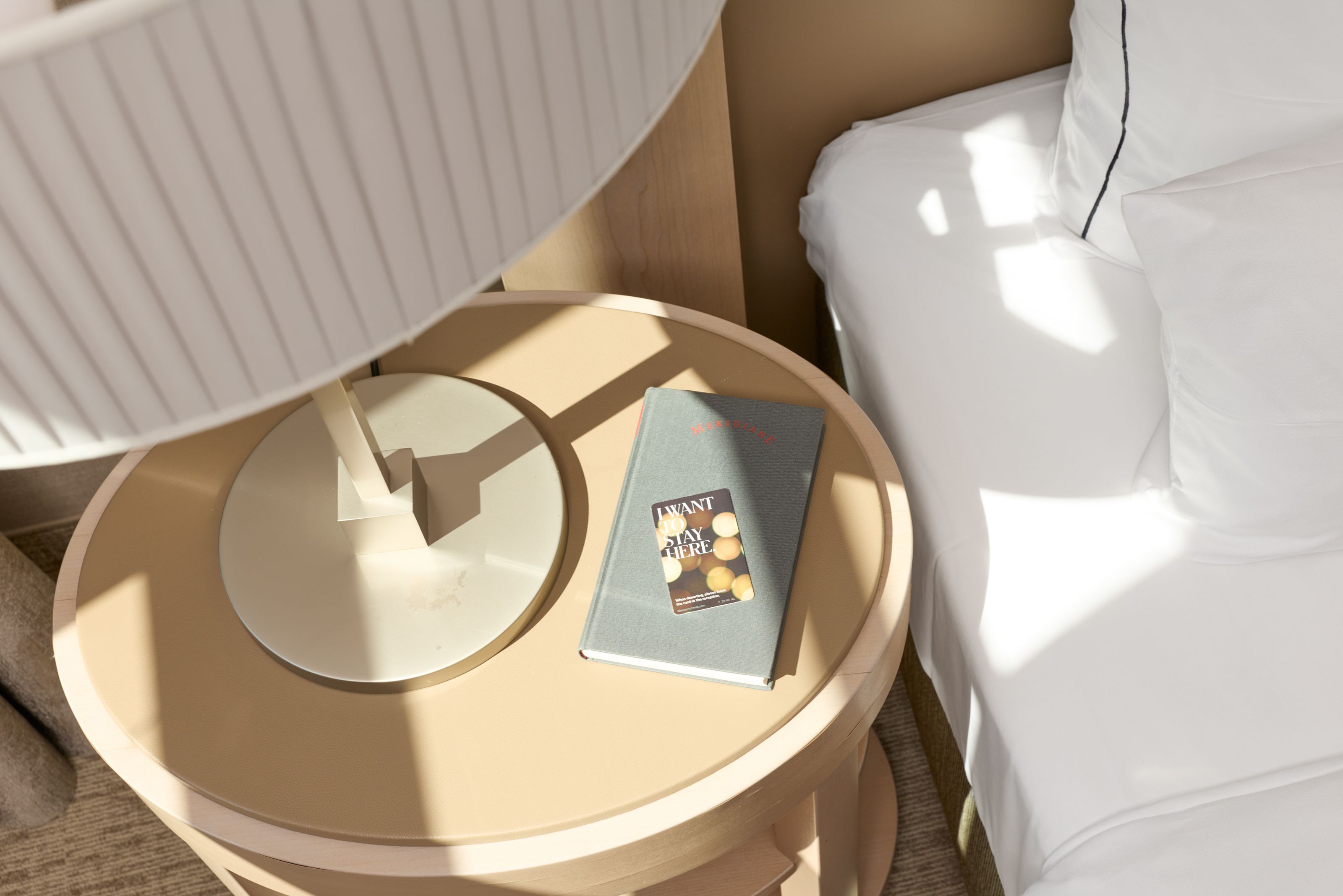 Beiger Nachttisch mit moderner Lampe und Buch neben einem weiß bezogenen Bett, Sonnenlicht fällt sanft auf die Möbel und erzeugt eine warme, einladende Atmosphäre.