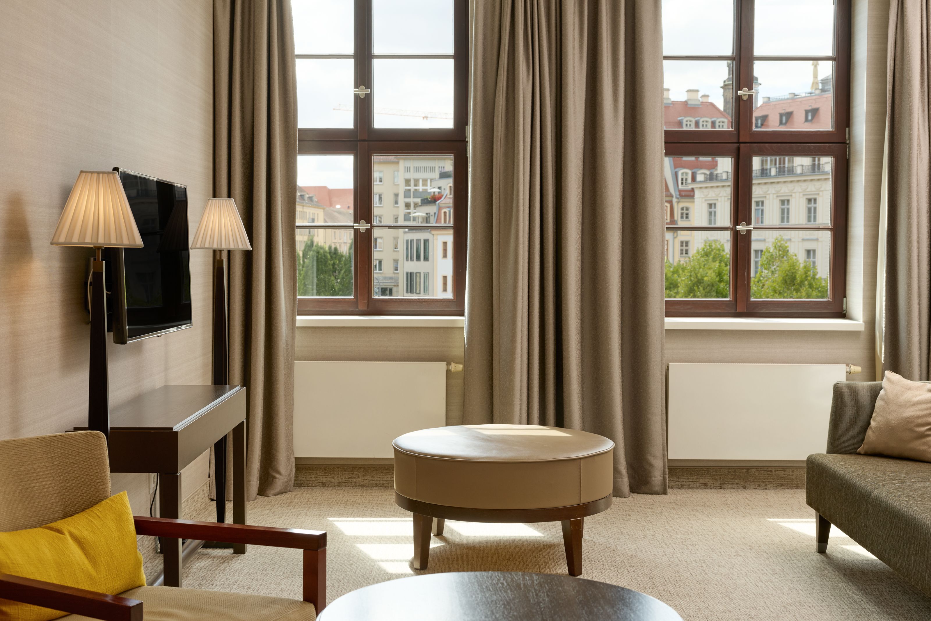 Helles Wohnzimmer mit großen Fenstern, neutralen Farben, bequemen Sitzmöbeln und Blick auf historische Gebäude. Gemütliche Atmosphäre für Entspannung und geselliges Beisammensein.