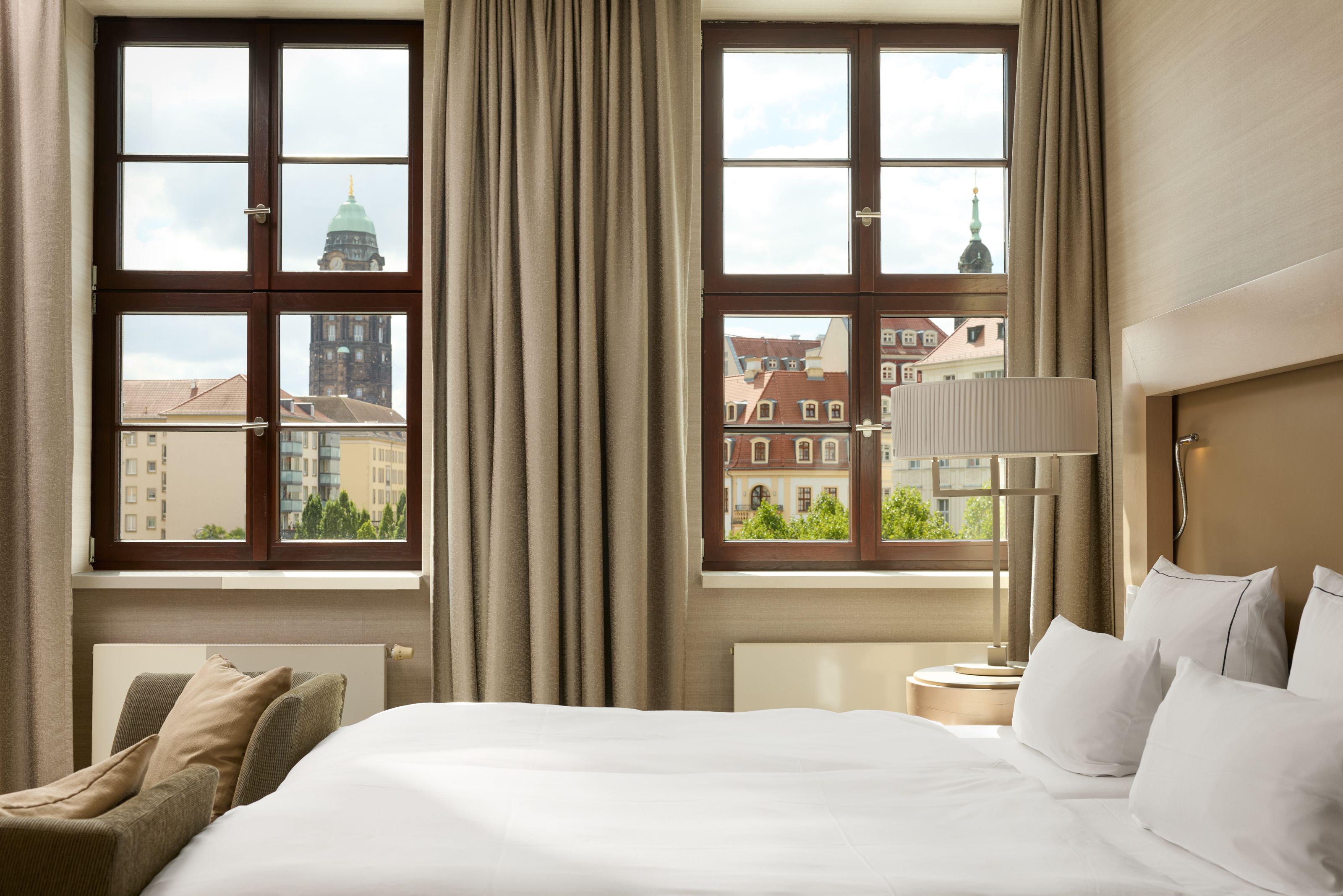 Helles Hotelzimmer mit großem Bett, gemütlichem Sessel und zwei Fenstern, die einen Blick auf historische Gebäude und Kirchtürme in der Stadt bieten.