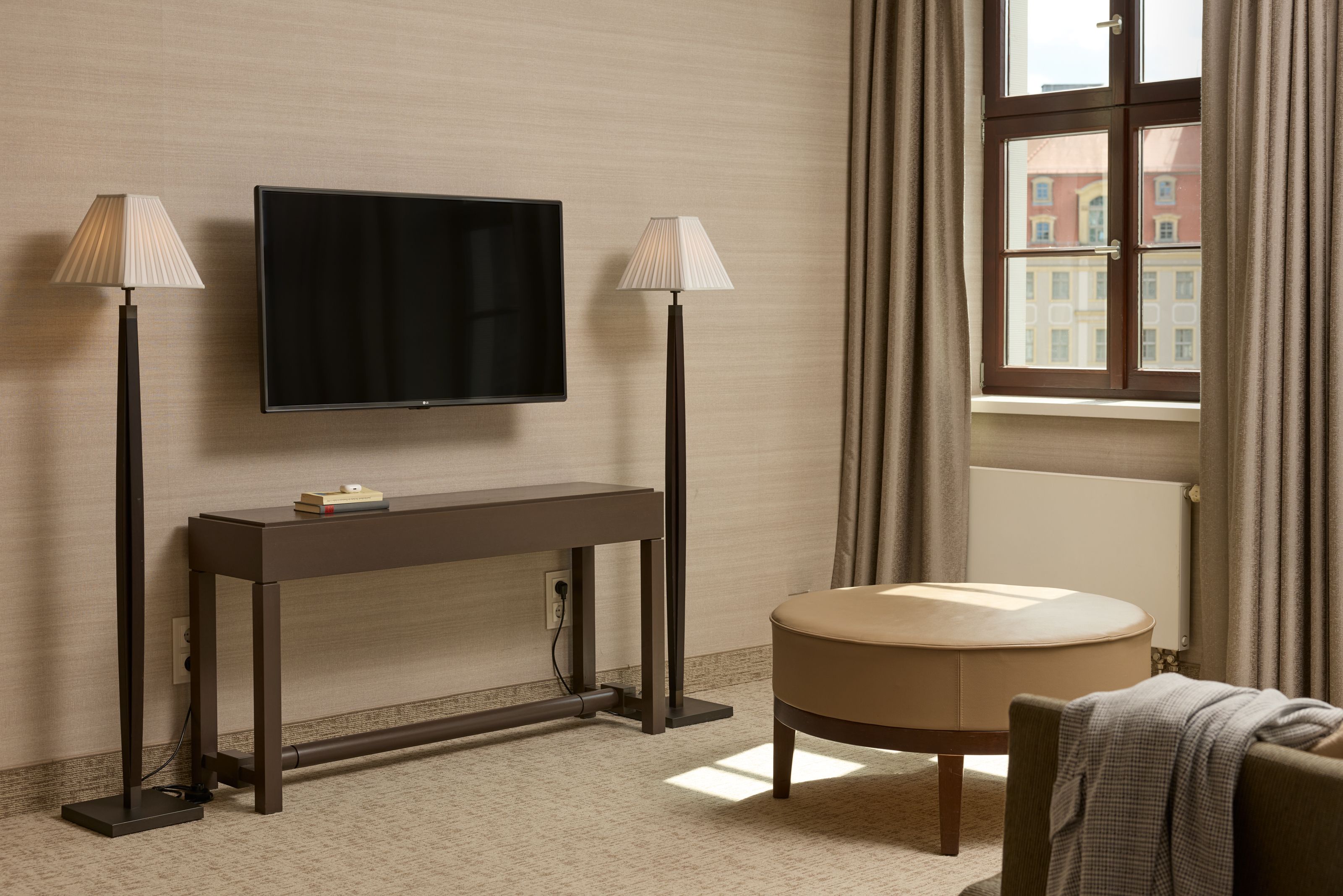 Heller, gemütlicher Wohnbereich mit großem Fenster, zwei Stehlampen, modernem Flachbildfernseher an der Wand, schmalem Konsolentisch und rundem, beigen Hocker, der weiches Licht und entspannte Atmosphäre vermittelt.