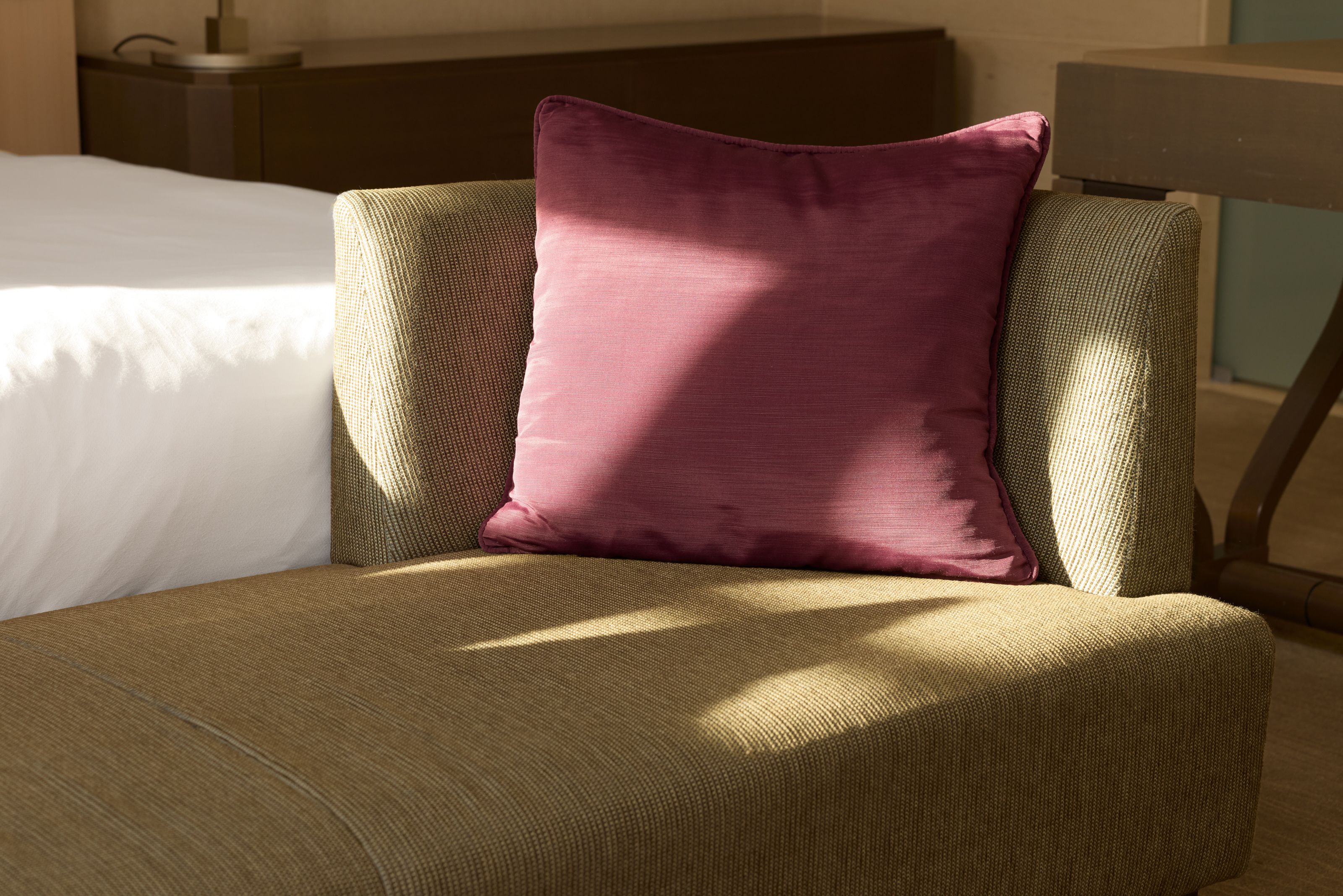 Gemütliche Sitzliege mit beigem Stoffbezug und einem kontrastierenden, weichen lila Kissen, Sonnenlicht fällt warm darauf, im Hintergrund ein weiß bezogenes Bett, die Szene strahlt Ruhe und Komfort aus.