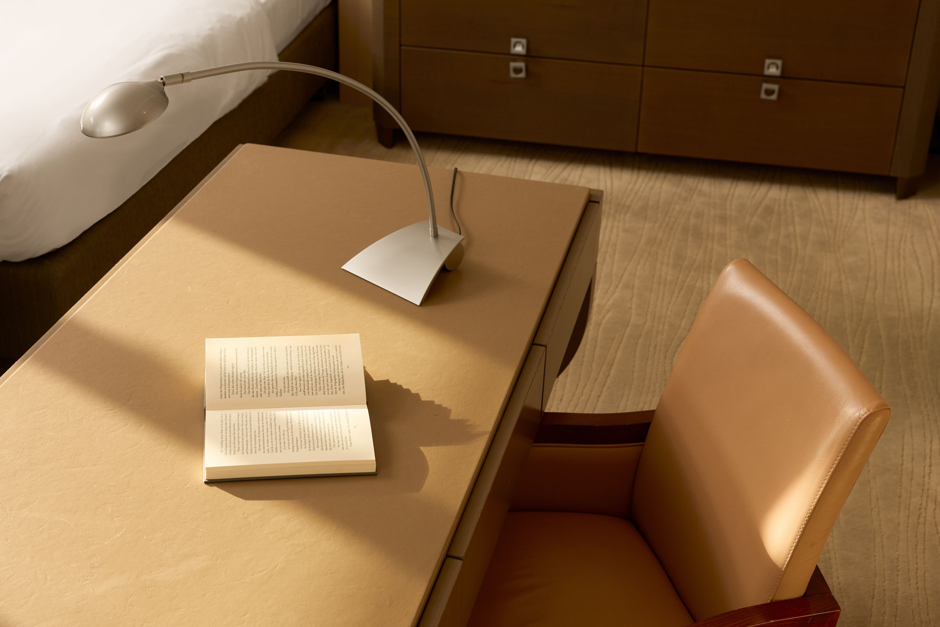 Schreibtisch in warmem Licht mit offenem Buch und moderner Leselampe, neben gemütlichem braunem Ledersessel, Teil eines stilvoll eingerichteten Hotelzimmers.