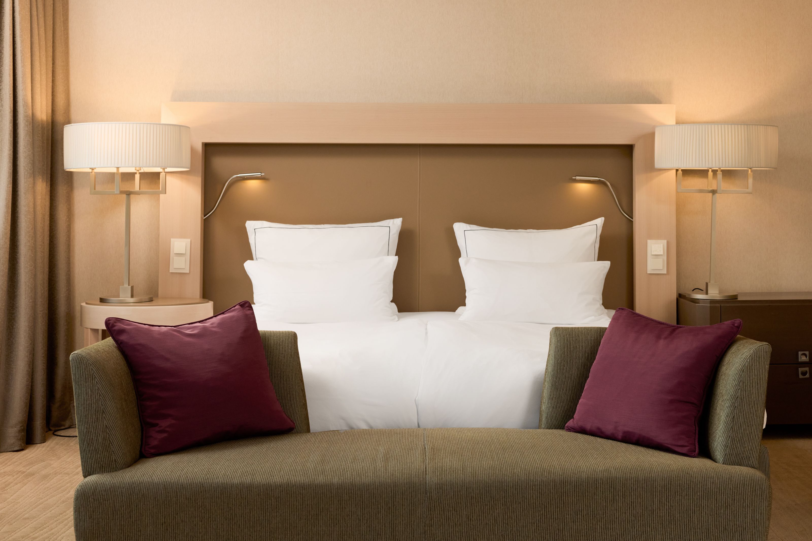 Helles Hotelzimmer mit einem großen Doppelbett, weißen Kissen, zwei Nachttischlampen und einem grauen Sofa mit zwei bordeauxroten Kissen davor, vermittelt Komfort und Ruhe für Gäste.