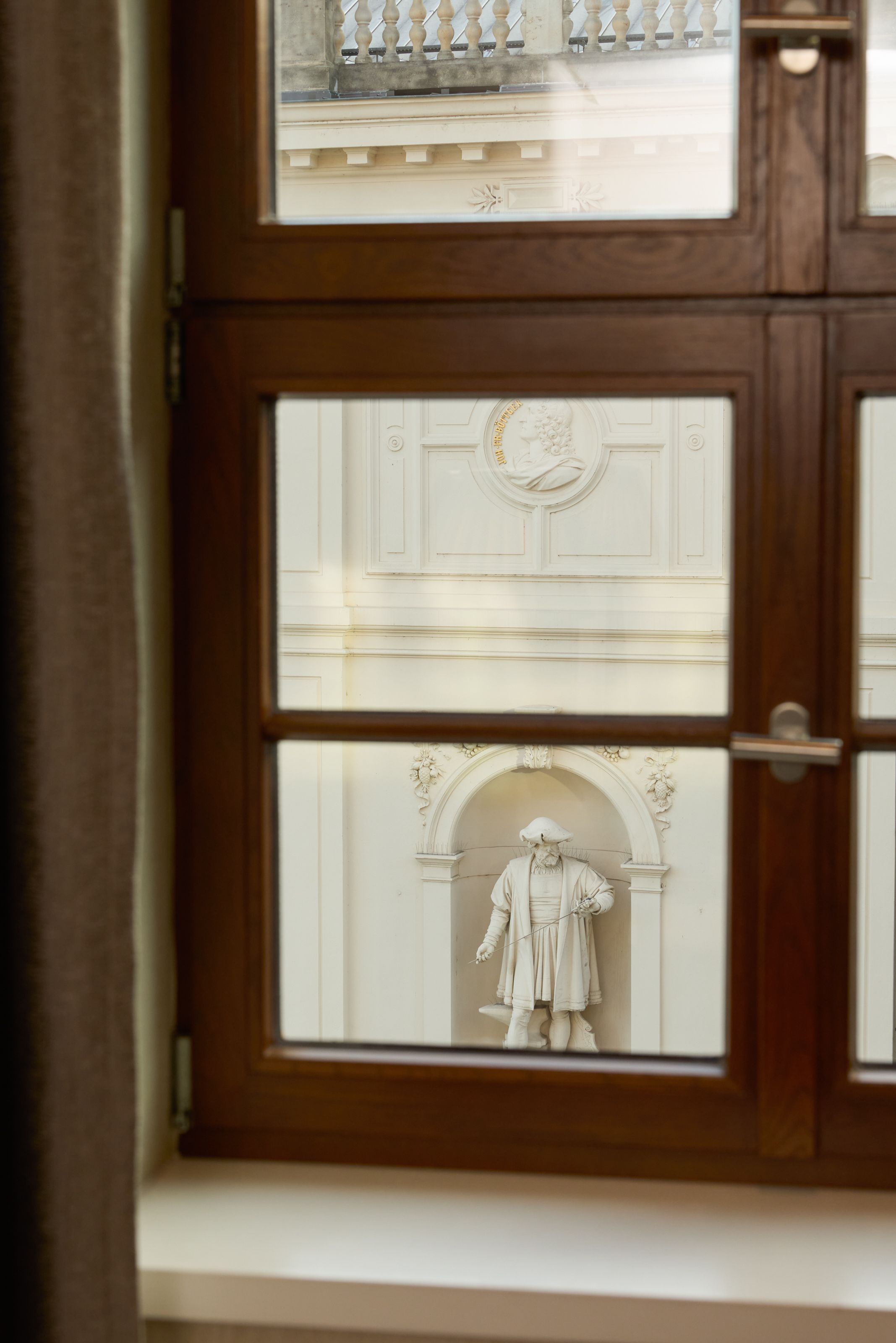 Ausblick durch ein Holzfenster auf eine weiße Fassade mit einer historischen Statue in einer Nische, die eine Figur in traditioneller Kleidung zeigt. Die Atmosphäre vermittelt historischen Charme und kulturelles Erbe.