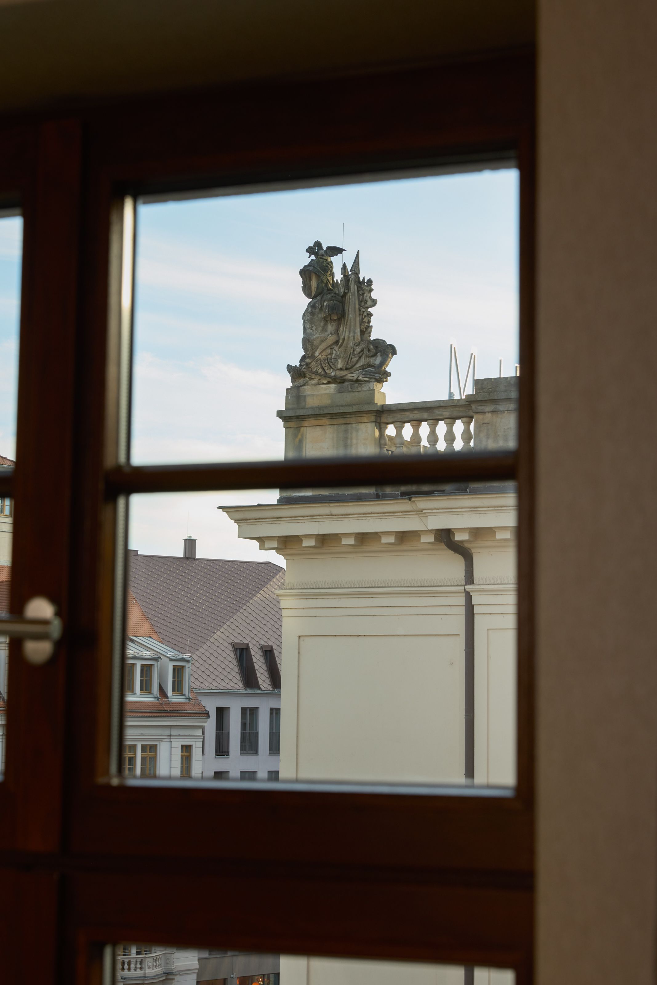 Blick aus einem Fenster auf ein historisches Gebäude mit einer kunstvollen Steinskulptur auf dem Dach, daneben sind Dächer weiterer Gebäude zu sehen. Der Himmel ist leicht bewölkt.
