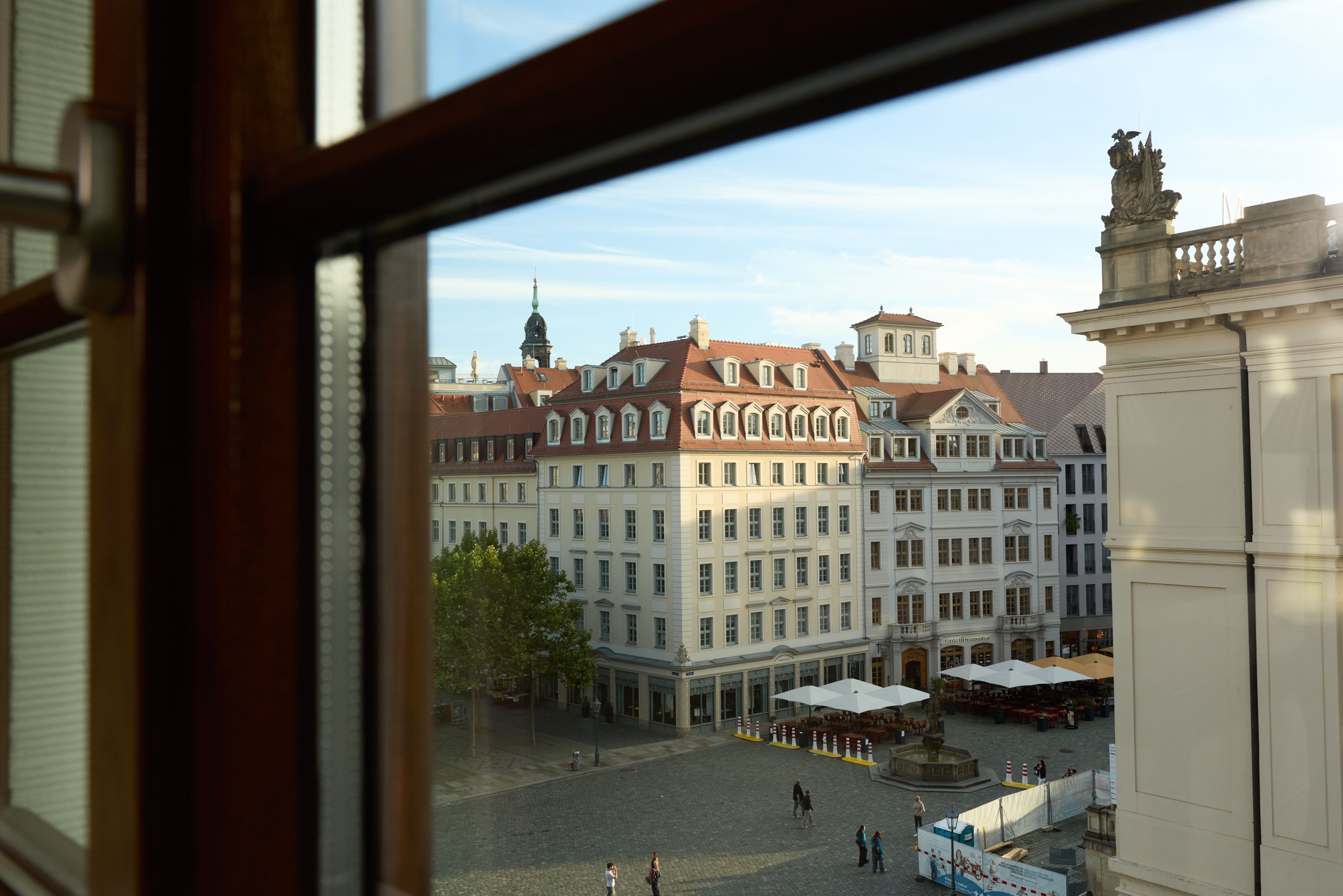 Blick aus einem Fenster auf einen historischen, sonnendurchfluteten Stadtplatz mit klassischen, mehrstöckigen Gebäuden und einem kleinen Café mit Außensitzplätzen. Der Platz wirkt ruhig, einige Menschen spazieren oder sitzen entspannt im Freien.