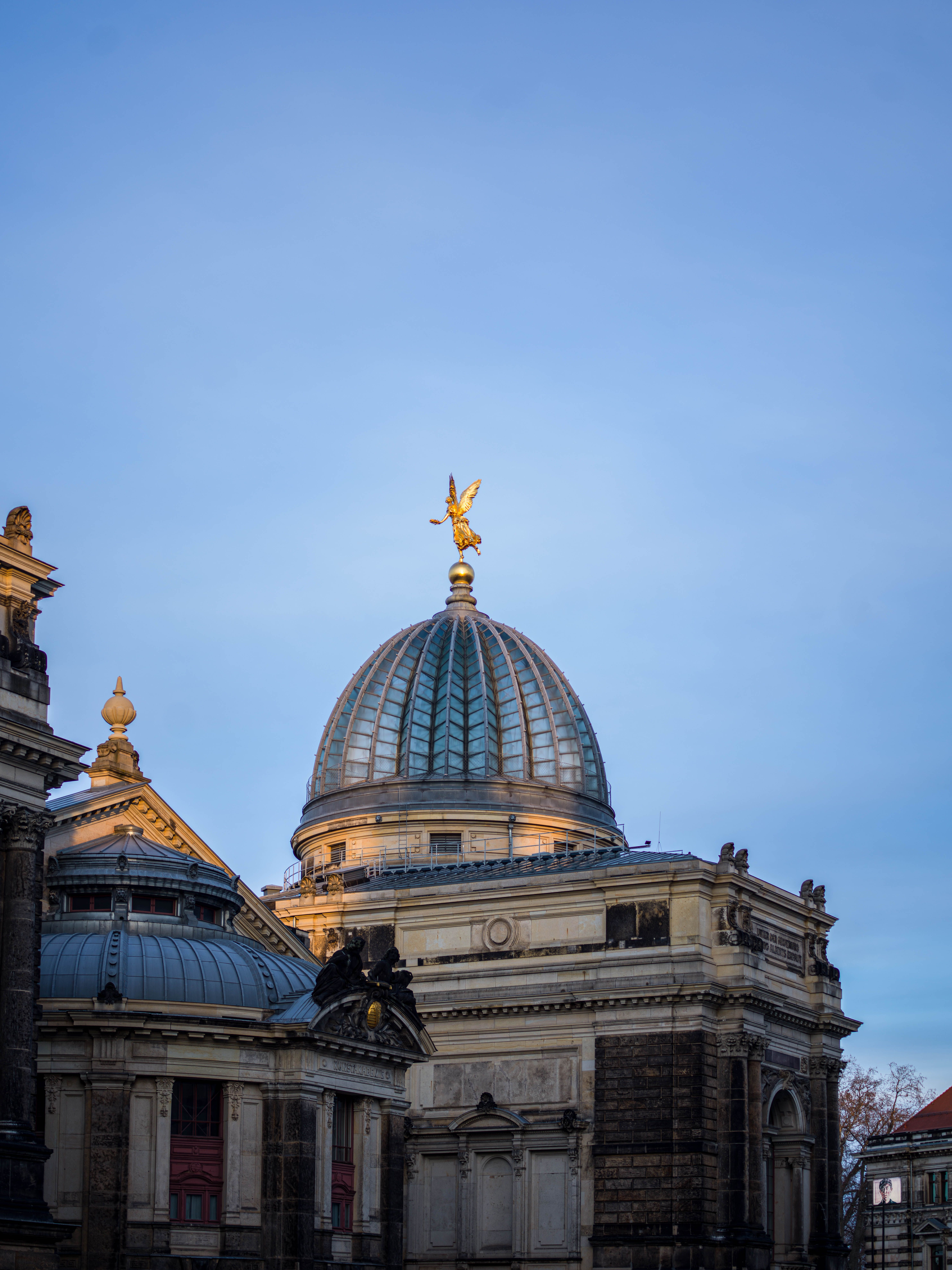 Kuppel eines historischen Gebäudes mit einer goldenen Engelsfigur auf der Spitze, vor klarem, blauem Himmel.
