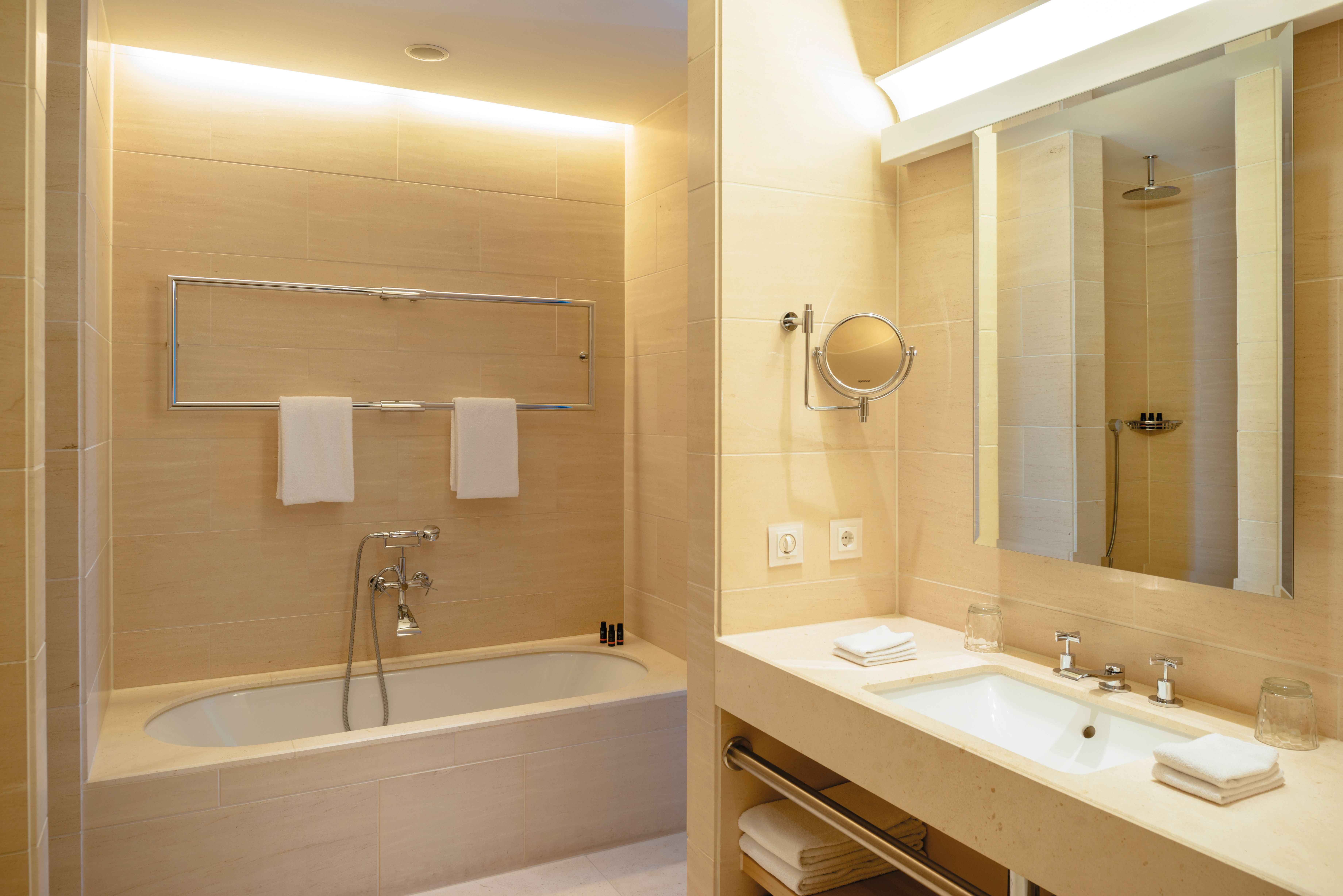 Modernes Badezimmer mit eingebauter Badewanne, hellem Waschbeckenbereich, großem Spiegel und Handtüchern. Ruhige, warme Beleuchtung schafft eine einladende Atmosphäre für Entspannung und Wohlbefinden.