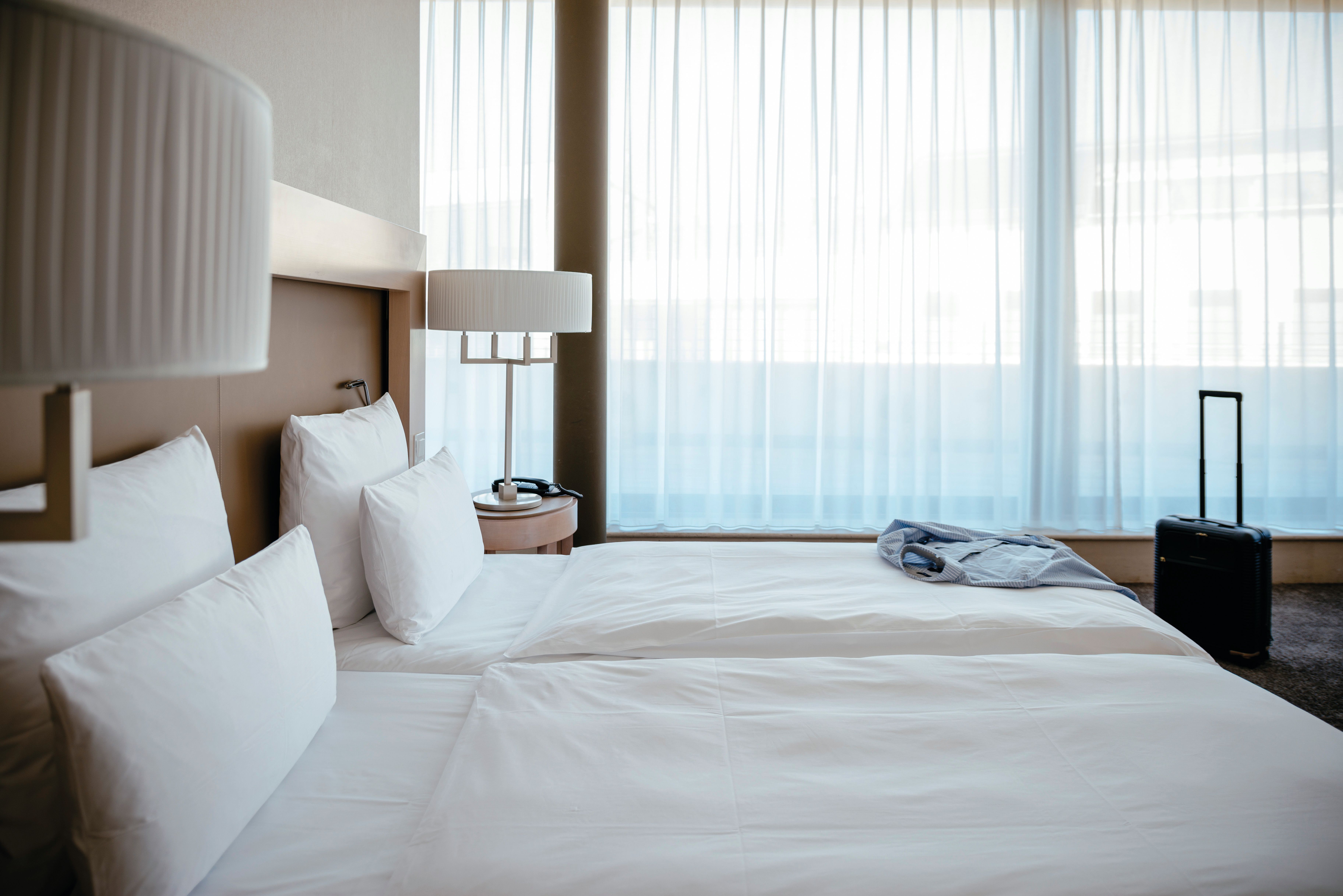 Helles Hotelzimmer mit großem Doppelbett, weißen Bettdecken und Kissen, sanfter Beleuchtung und bodentiefen Fenstern, die Tageslicht hereinlassen. Koffer und Jacke liegen für die Ankunft bereit.