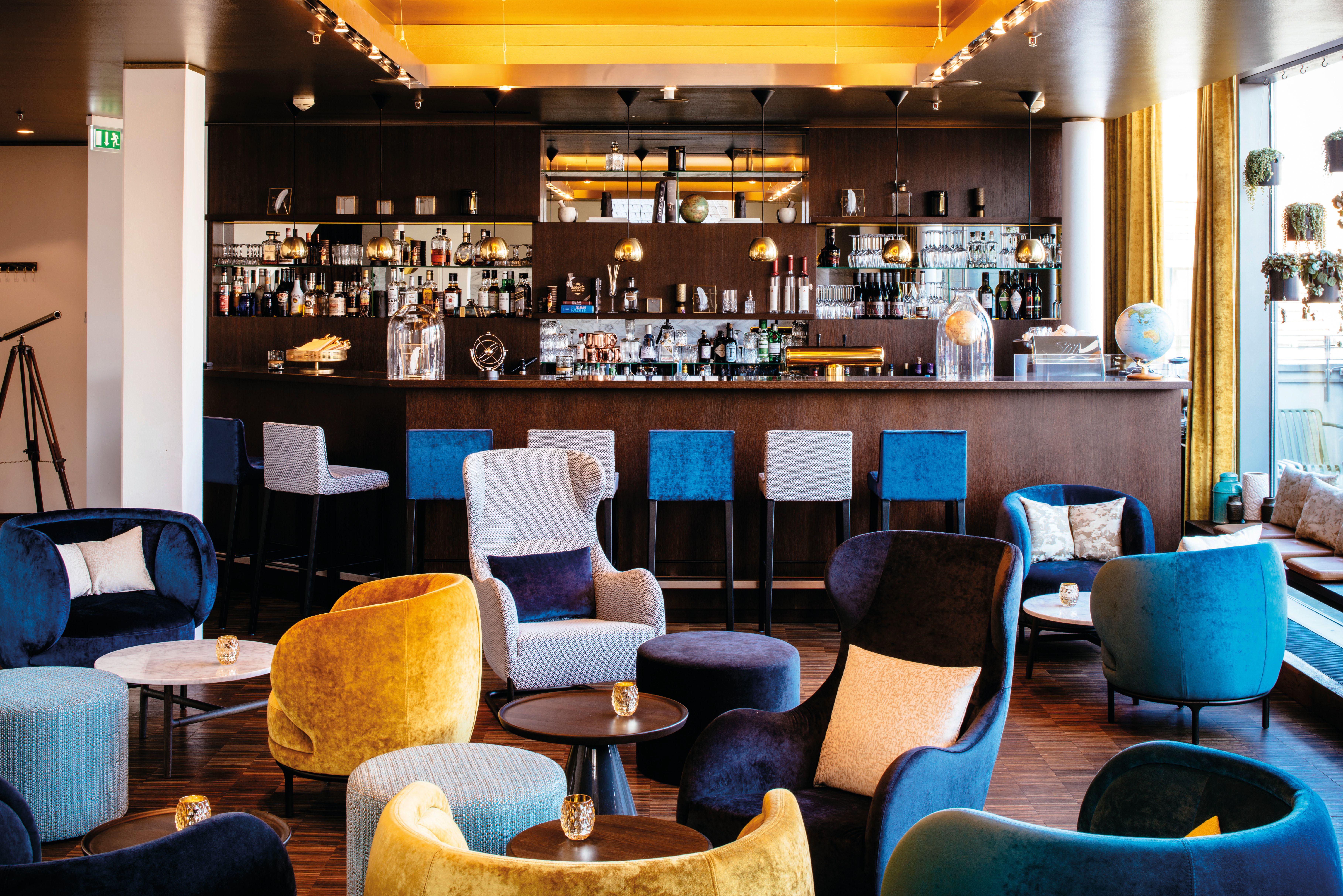 Elegante Hotelbar mit gemütlichen Sesseln in Blau und Gelb, dekorativen Kissen und kleinen Tischen. Große Fenster lassen Tageslicht herein und schaffen eine einladende Atmosphäre zum Entspannen und Genießen von Getränken.