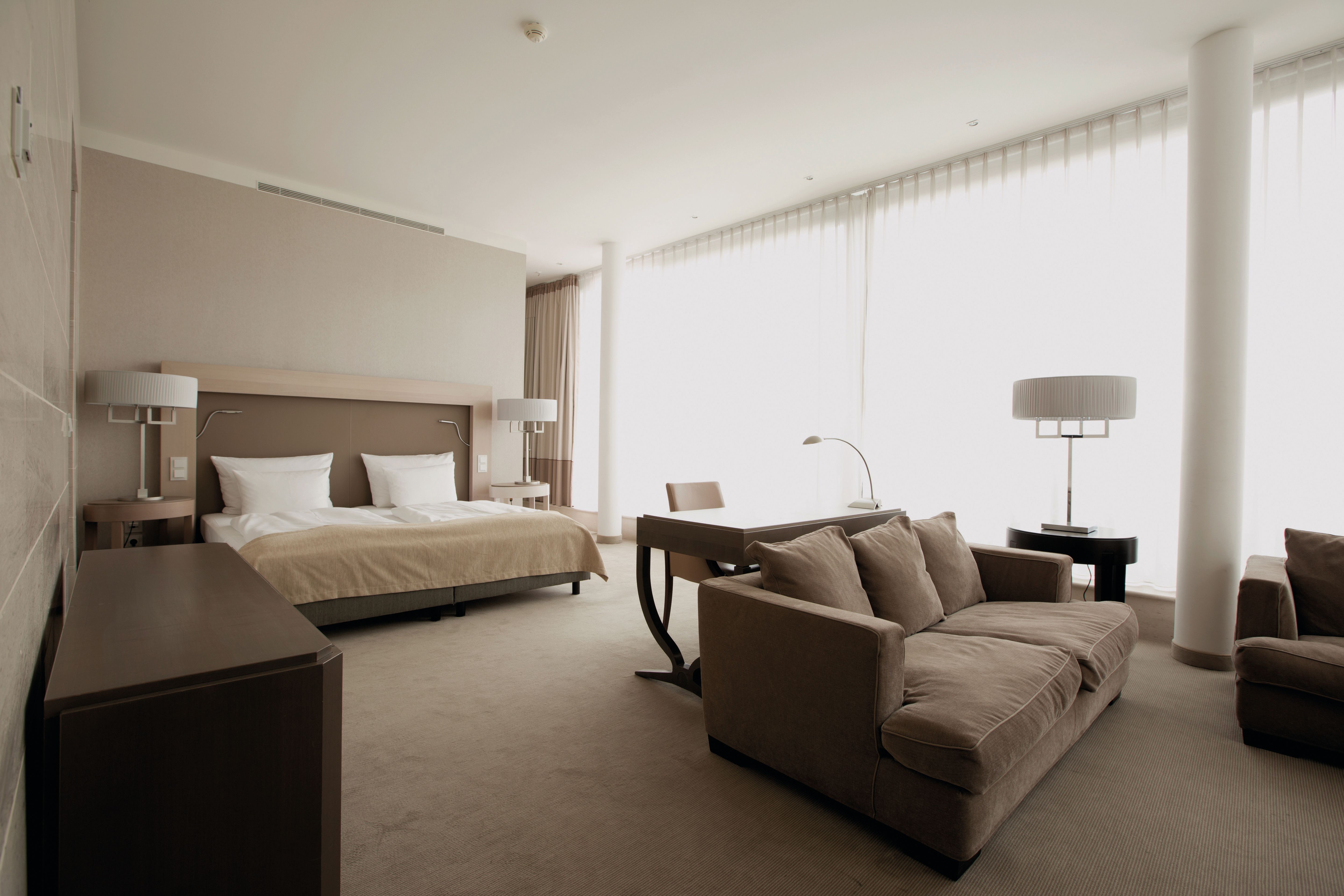 Helles, modern eingerichtetes Hotelzimmer mit großem Doppelbett, gemütlichem Sofa und Schreibtisch vor bodentiefen Fenstern, die viel Tageslicht hereinlassen.