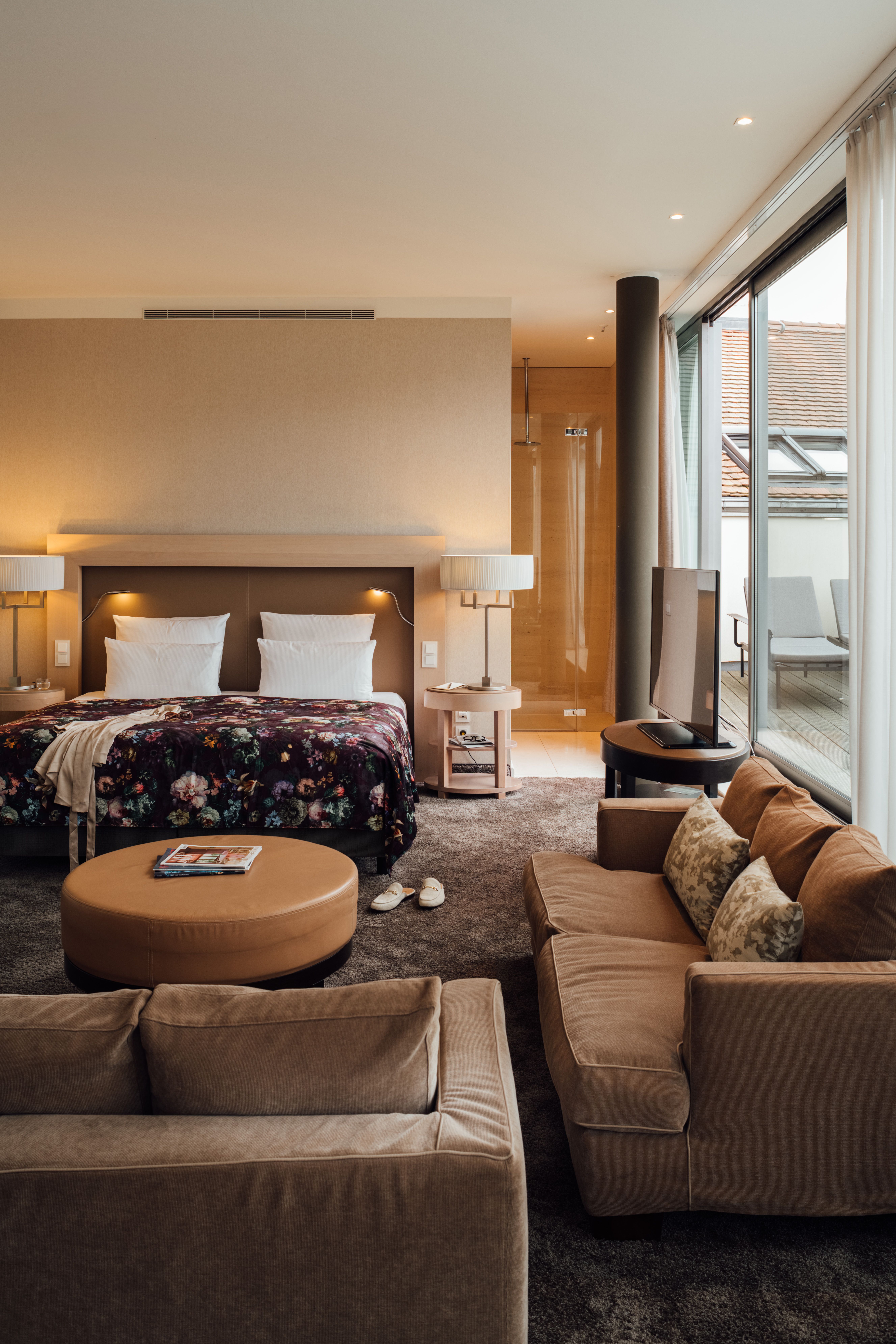 Komfortables Hotelzimmer mit Doppelbett, floral gemustertem Überwurf, Sitzbereich mit Sofa und Sessel sowie großem Fenster mit Zugang zu Balkon und Blick auf Dächer. Warme, einladende Beleuchtung für entspannte Atmosphäre.