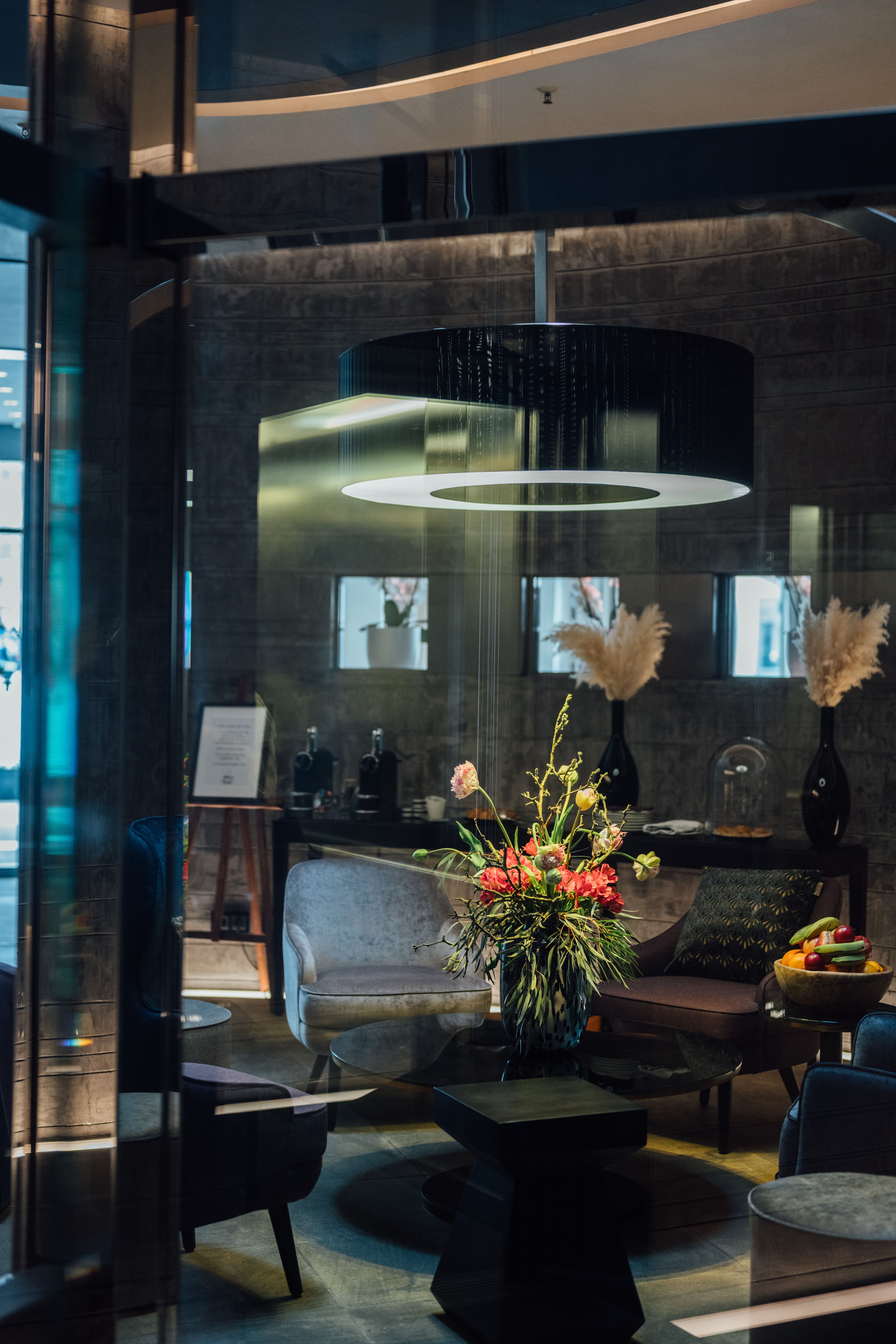 Moderner Hotel-Loungebereich mit stilvollen Sitzmöbeln, dunklem Ambiente und einem großen, runden Deckenleuchter. Auf dem Tisch steht ein bunter Blumenstrauß, im Hintergrund dekorative Vasen und entspanntes, gemütliches Flair.