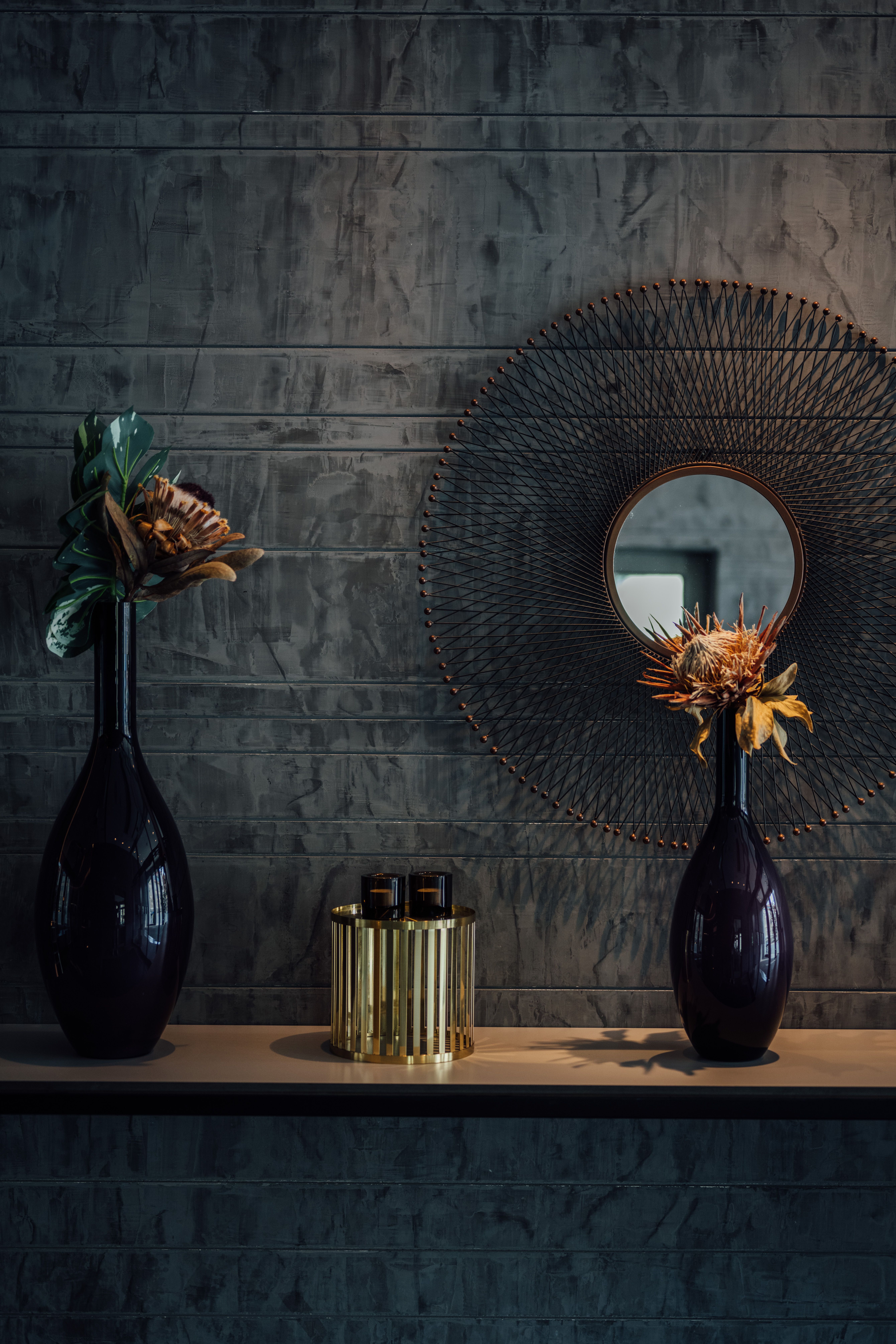 Dunkles, stilvolles Interieur mit zwei hohen Vasen, darin dekorative Blumen, und rundem, kunstvollem Spiegel an der Wand. Stimmungsvoll und elegant, ideal für ein hochwertiges Hotelambiente.