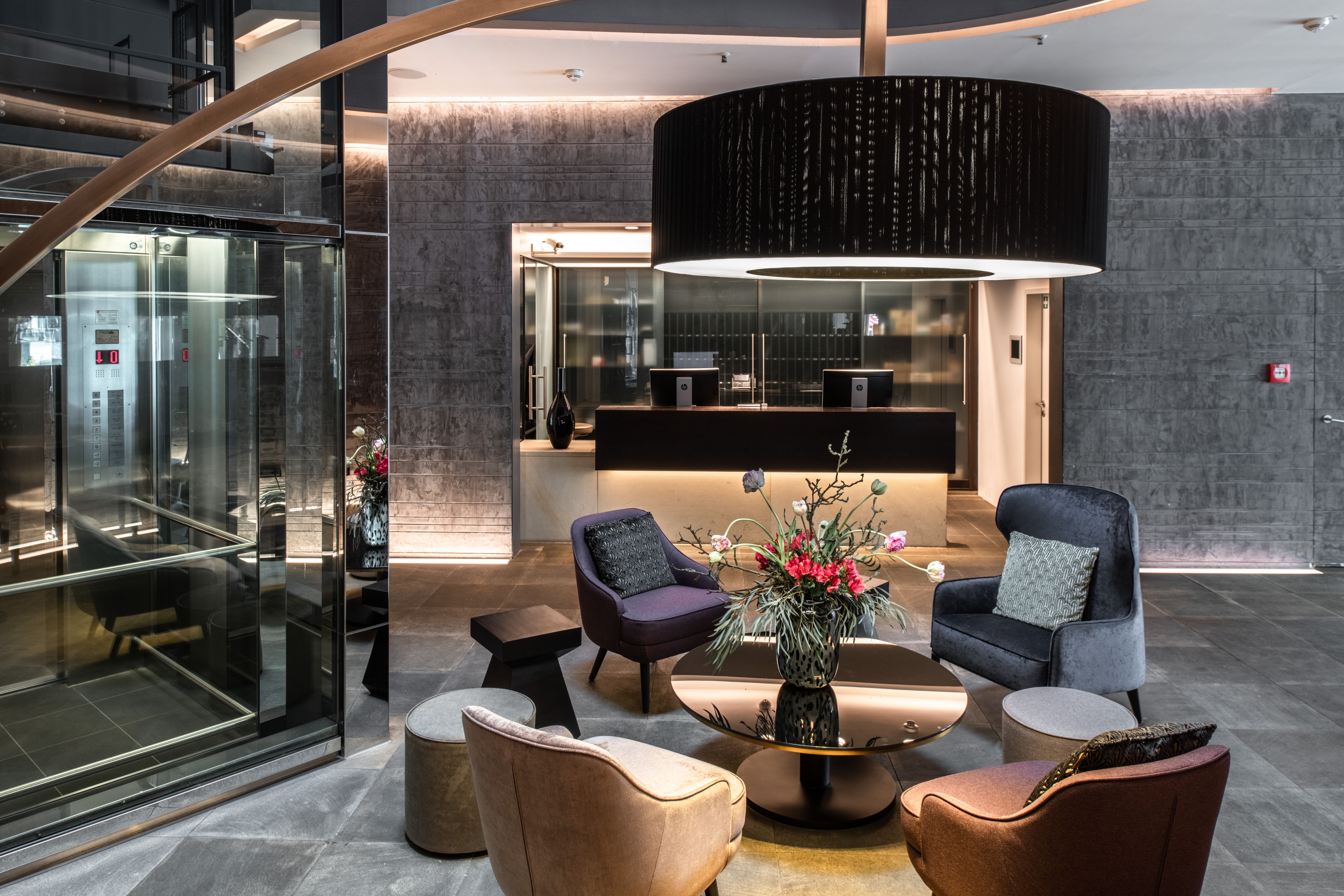 Moderner Hotel-Loungebereich mit gemütlichen Sesseln um einen runden Glastisch, dekoriert mit frischen Blumen. Sanfte Beleuchtung sorgt für ein einladendes und entspanntes Ambiente. Heller Empfangsbereich mit Aufzug in der Nähe.