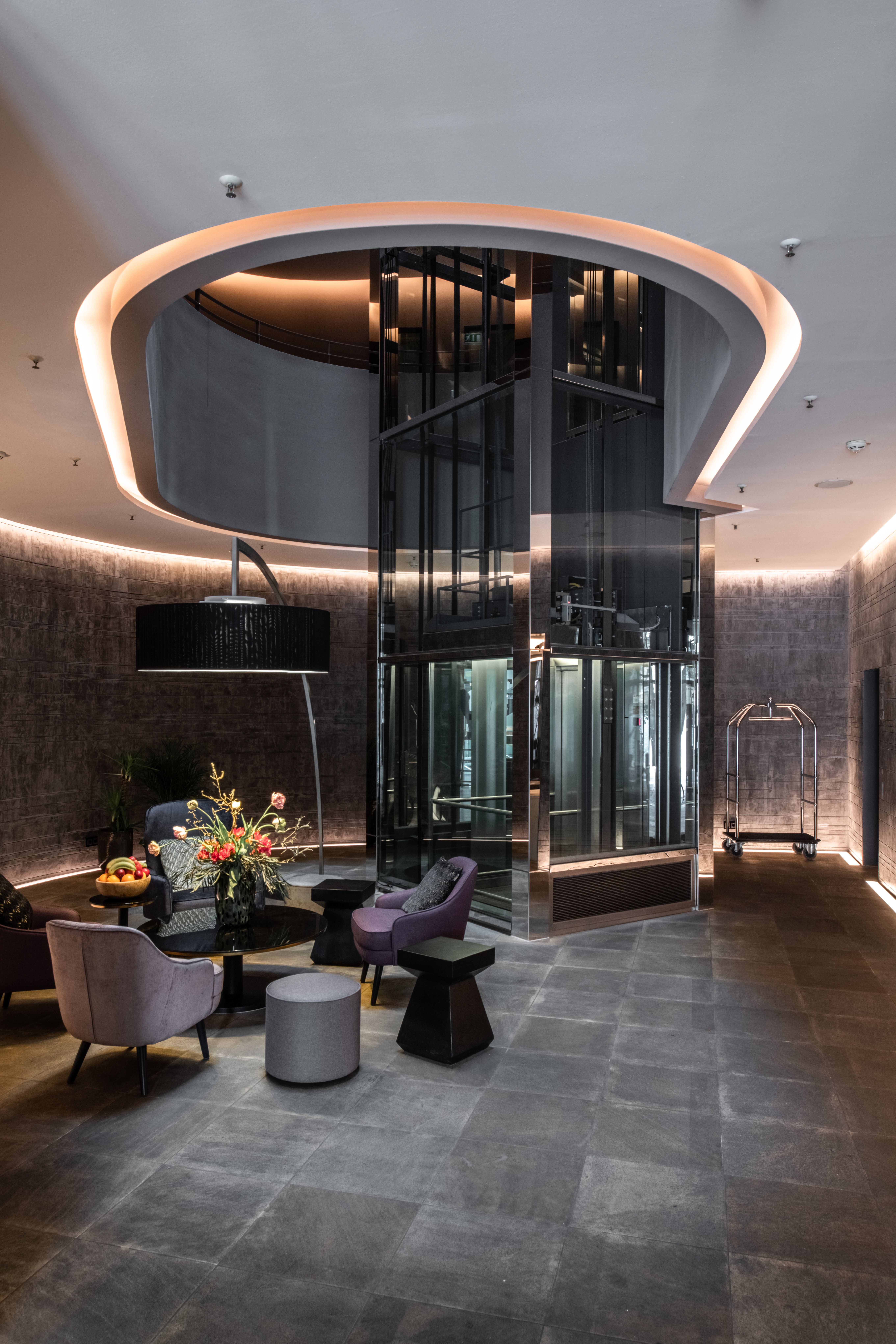 Moderner Hotellobby mit stilvoller Beleuchtung, gemütlichen Sesseln und Glaselementen. Zentrale Glasfahrstuhlanlage unter einem beleuchteten Rundbogen. Elegantes, einladendes Ambiente für Gäste zum Entspannen.