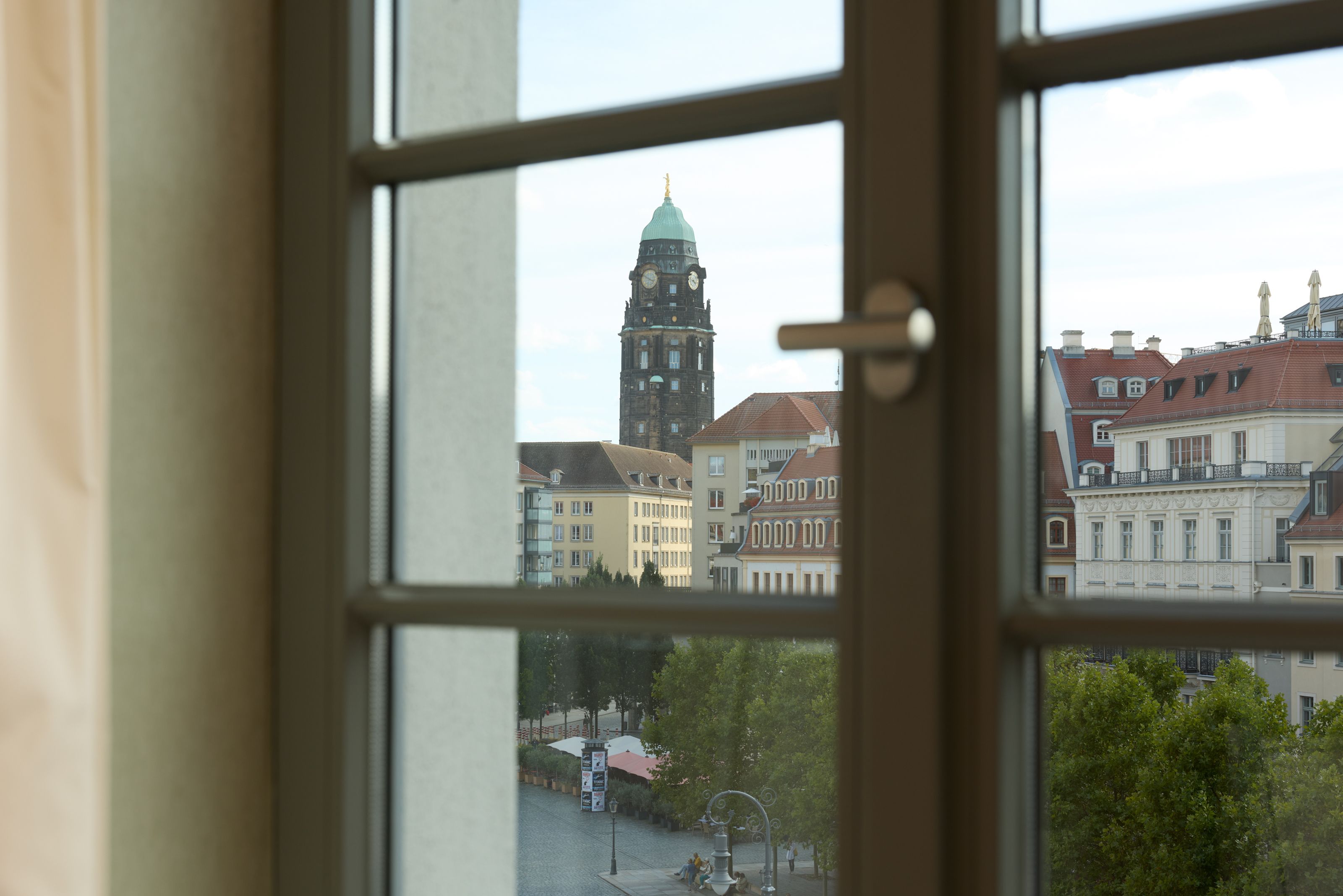 The Cloud One Dresden-Frauenkirche Blick aus einem Hotelzimmerfenster auf eine historische Stadt mit einem hohen Kirchturm und klassischer Architektur, umgeben von Bäumen und einer ruhigen Straße. Die Atmosphäre wirkt friedlich und einladend.