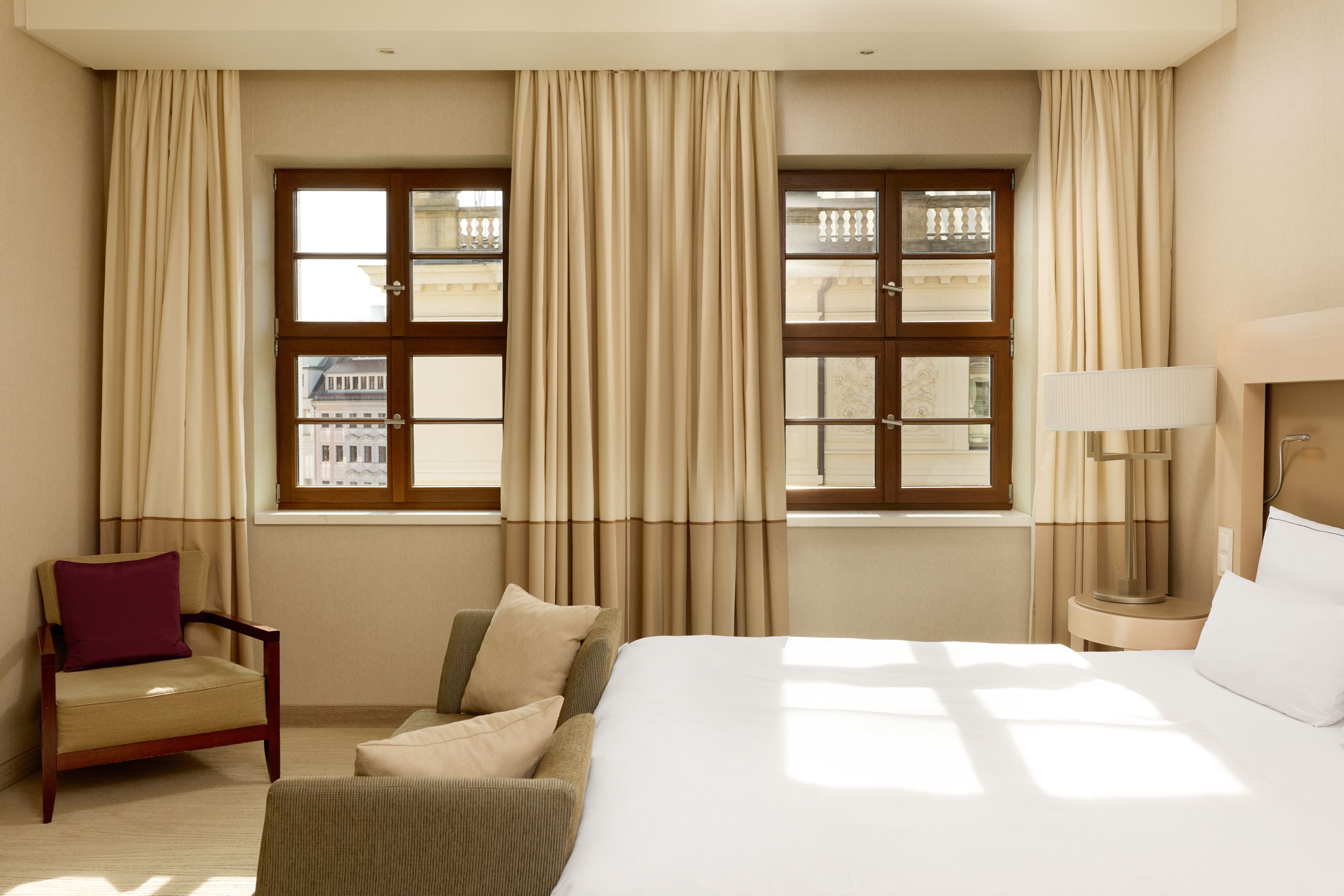 The Cloud One Dresden-Frauenkirche Helles, gemütlich eingerichtetes Hotelzimmer mit großem Bett, Sofa und Sesseln, zwei großen Fenstern mit Blick auf ein klassisches Stadtgebäude. Natürlicher Sonnenschein schafft eine warme und einladende Atmosphäre.