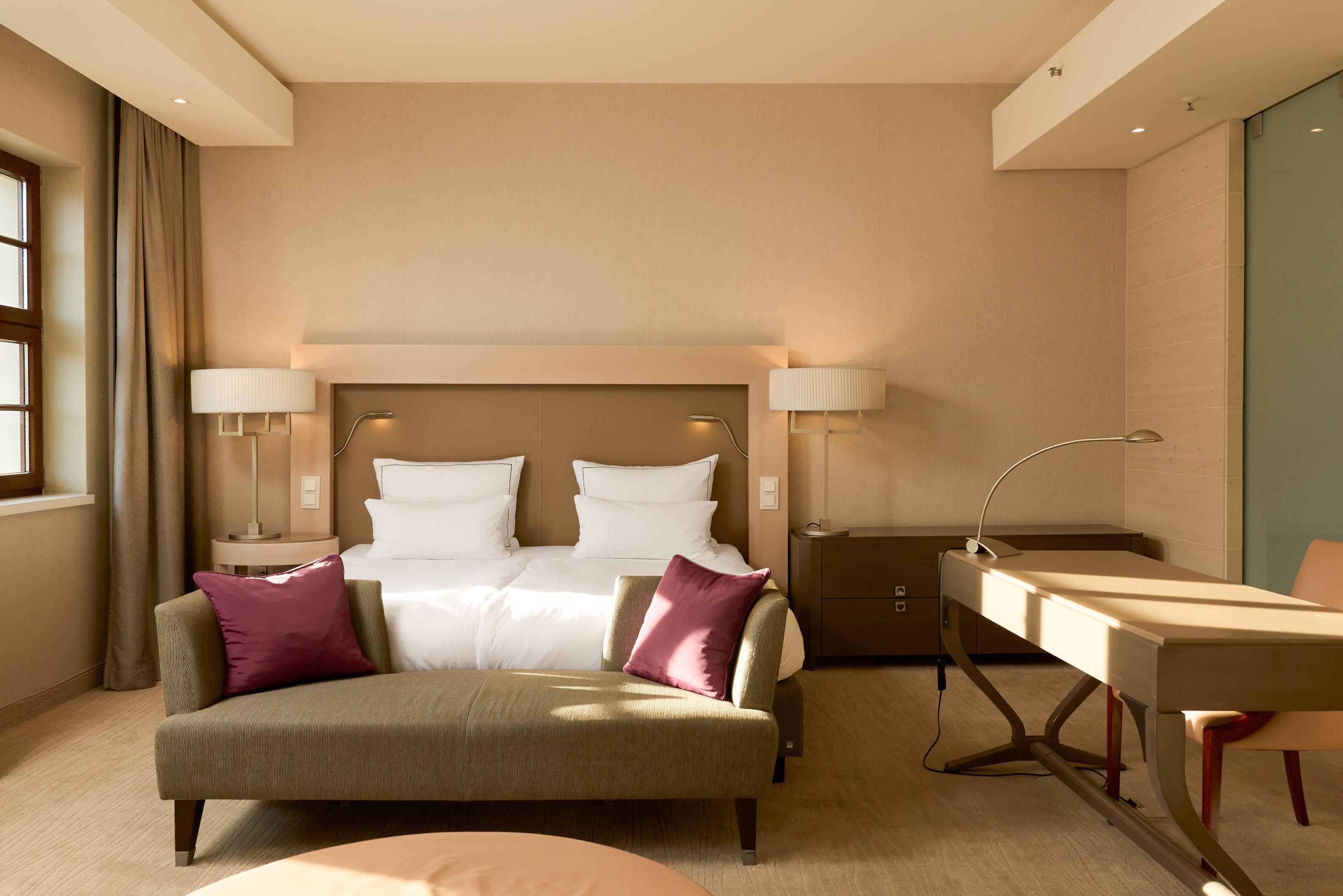 The Cloud One Dresden-Frauenkirche Elegantes Hotelzimmer mit großem Doppelbett, weichen Kissen und zwei gemütlichen Sesseln mit beerenfarbenen Kissen. Helle, warme Beleuchtung und Fenster mit Blick nach draußen schaffen eine entspannte Atmosphäre für Erholung und Arbeitskomfort.