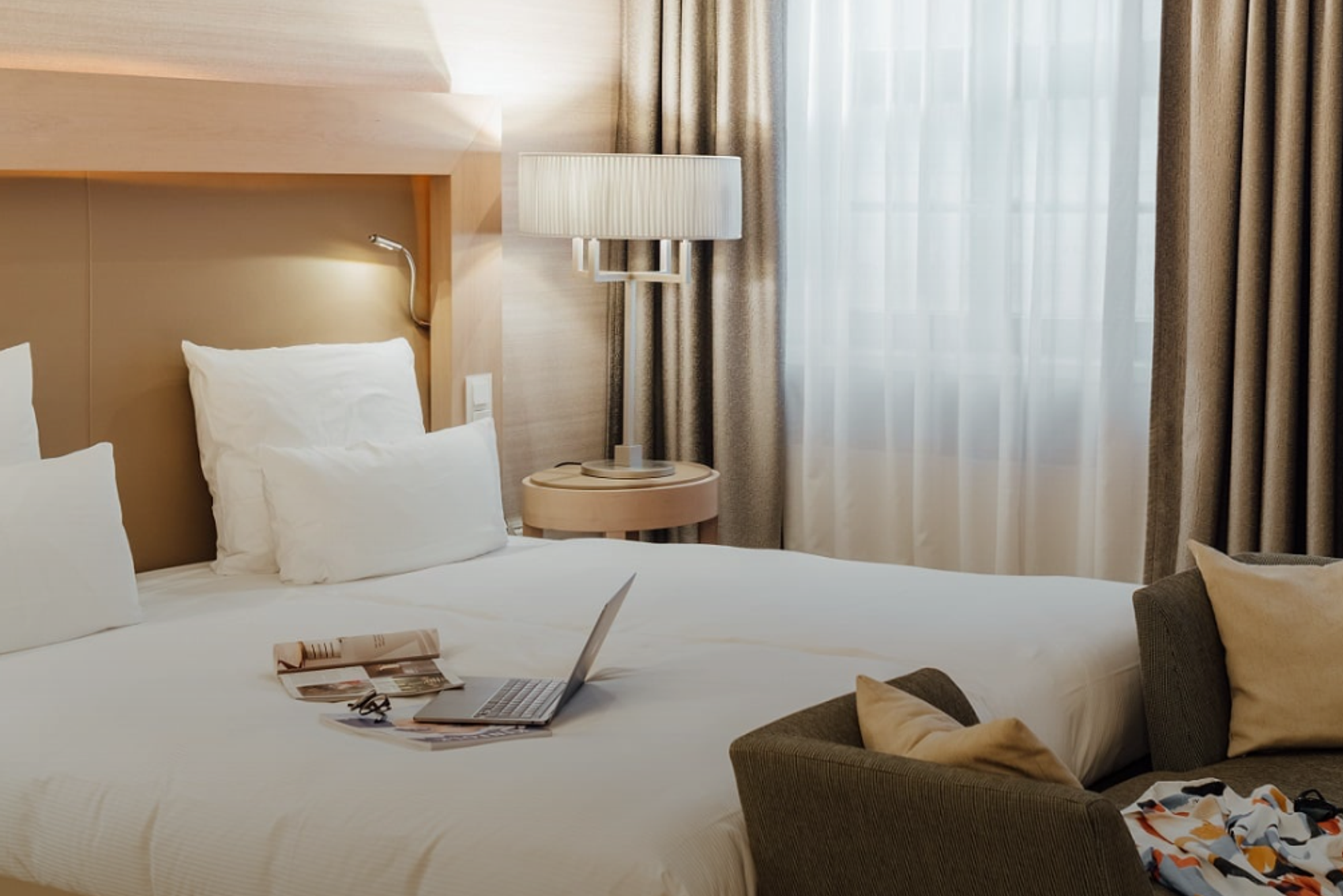 Helles, modern eingerichtetes Hotelzimmer mit großem Bett, weichen Kissen und Laptop auf der Bettdecke. Sanfte Beleuchtung und Vorhänge schaffen eine gemütliche Atmosphäre, ergänzt durch einen bequemen Sessel mit Kissen. Ideal für entspannte Aufenthalte oder Arbeit im Zimmer.