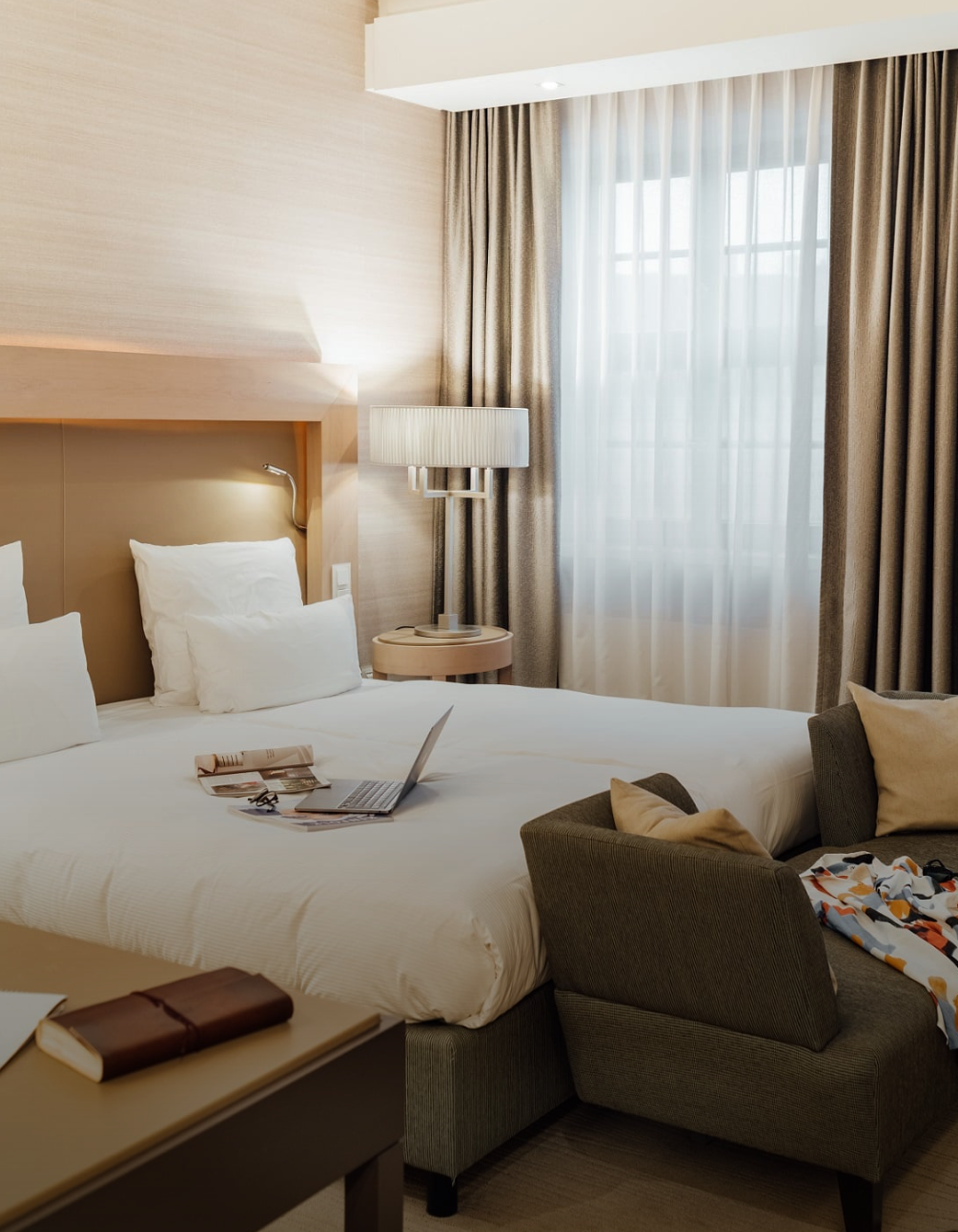 Helles, modern eingerichtetes Hotelzimmer mit großem Bett, weichen Kissen und Laptop auf der Bettdecke. Sanfte Beleuchtung und Vorhänge schaffen eine gemütliche Atmosphäre, ergänzt durch einen bequemen Sessel mit Kissen. Ideal für entspannte Aufenthalte oder Arbeit im Zimmer. Helles, modern eingerichtetes Hotelzimmer mit großem Bett, weichen Kissen und Laptop auf der Bettdecke. Sanfte Beleuchtung und Vorhänge schaffen eine gemütliche Atmosphäre, ergänzt durch einen bequemen Sessel mit Kissen. Ideal für entspannte Aufenthalte oder Arbeit im Zimmer.