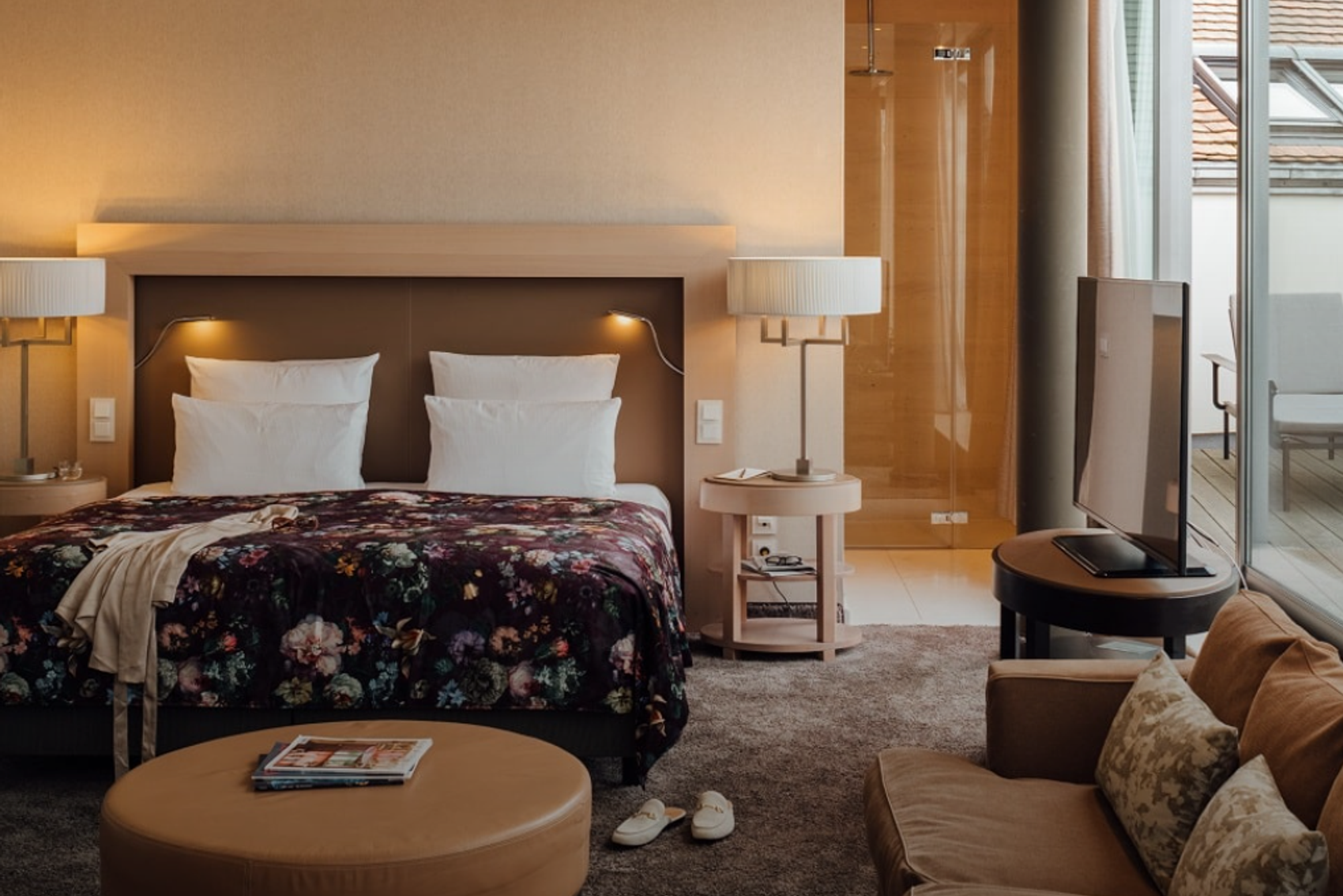 Modernes Hotelzimmer mit großem Doppelbett, bequemen Sofas und bodentiefen Fenstern, die viel Tageslicht und Blick auf ein Dach bieten. Gemütliche Atmosphäre mit warmen Farben und elegantem Interieur.