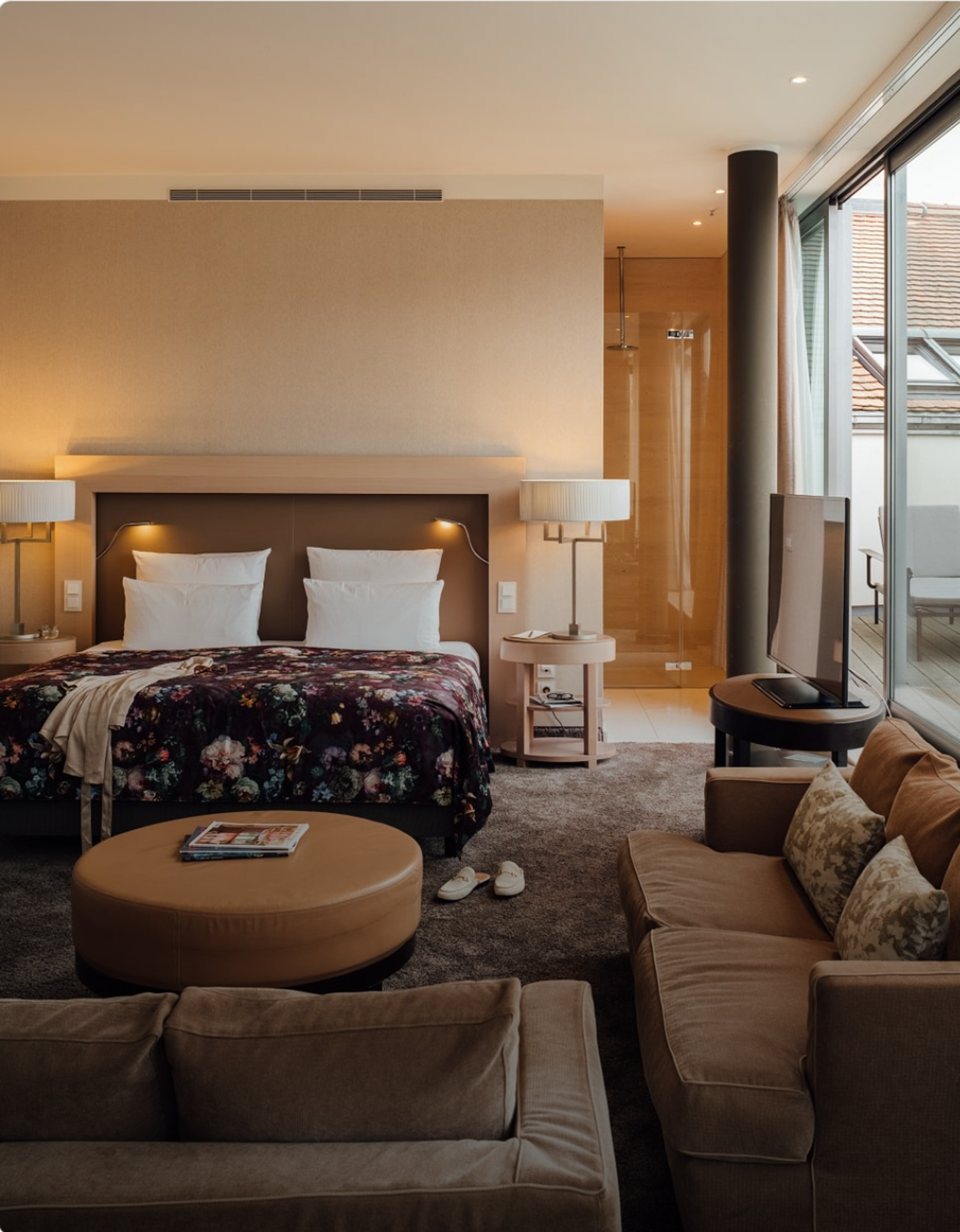 Modernes Hotelzimmer mit großem Doppelbett, bequemen Sofas und bodentiefen Fenstern, die viel Tageslicht und Blick auf ein Dach bieten. Gemütliche Atmosphäre mit warmen Farben und elegantem Interieur. Modernes Hotelzimmer mit großem Doppelbett, bequemen Sofas und bodentiefen Fenstern, die viel Tageslicht und Blick auf ein Dach bieten. Gemütliche Atmosphäre mit warmen Farben und elegantem Interieur.