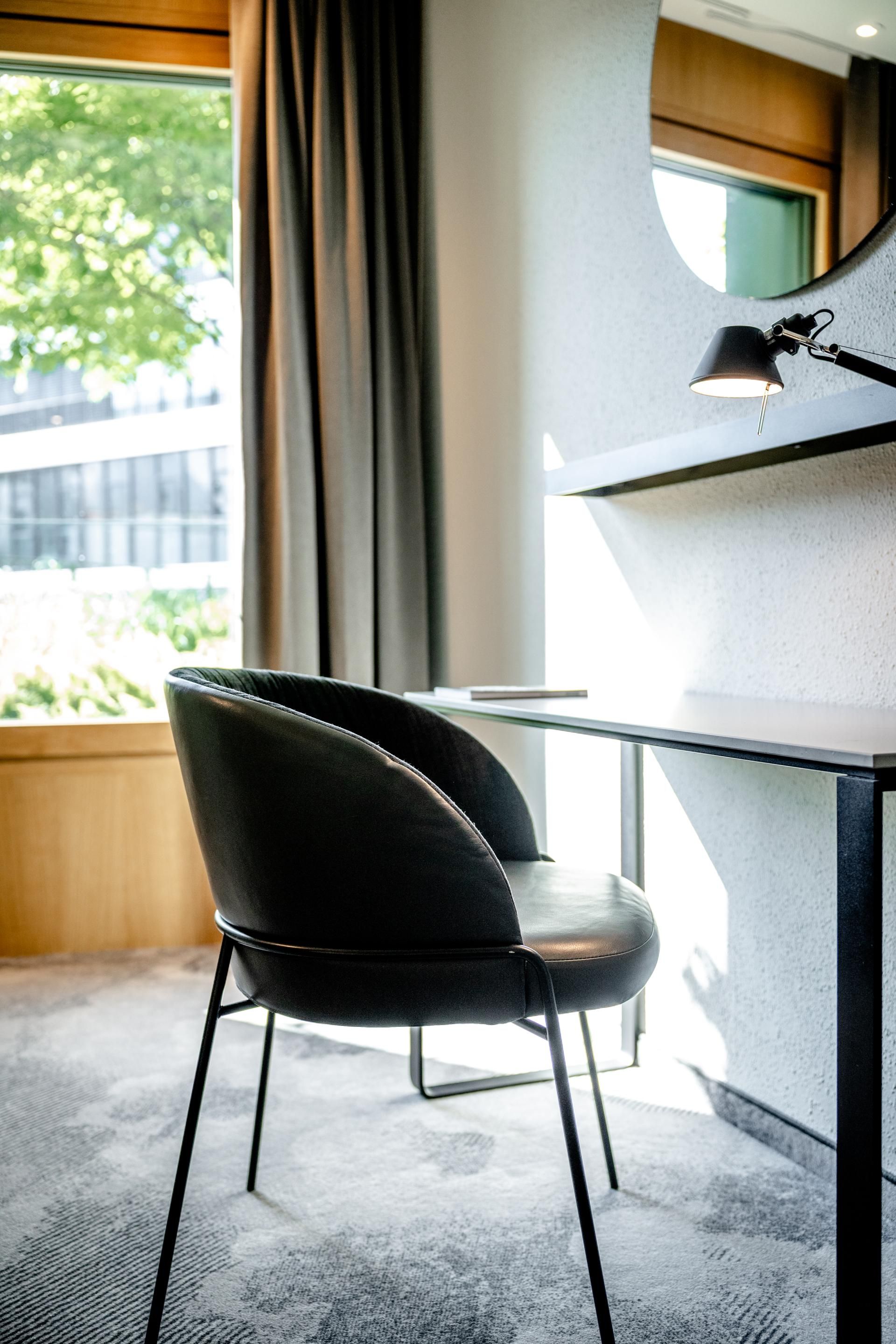 Moderner Arbeitsplatz mit schwarzem Lederstuhl, schlichter Schreibtisch und Wandleuchte, natürliches Licht durch großen Fensterbereich und Blick ins Grüne.