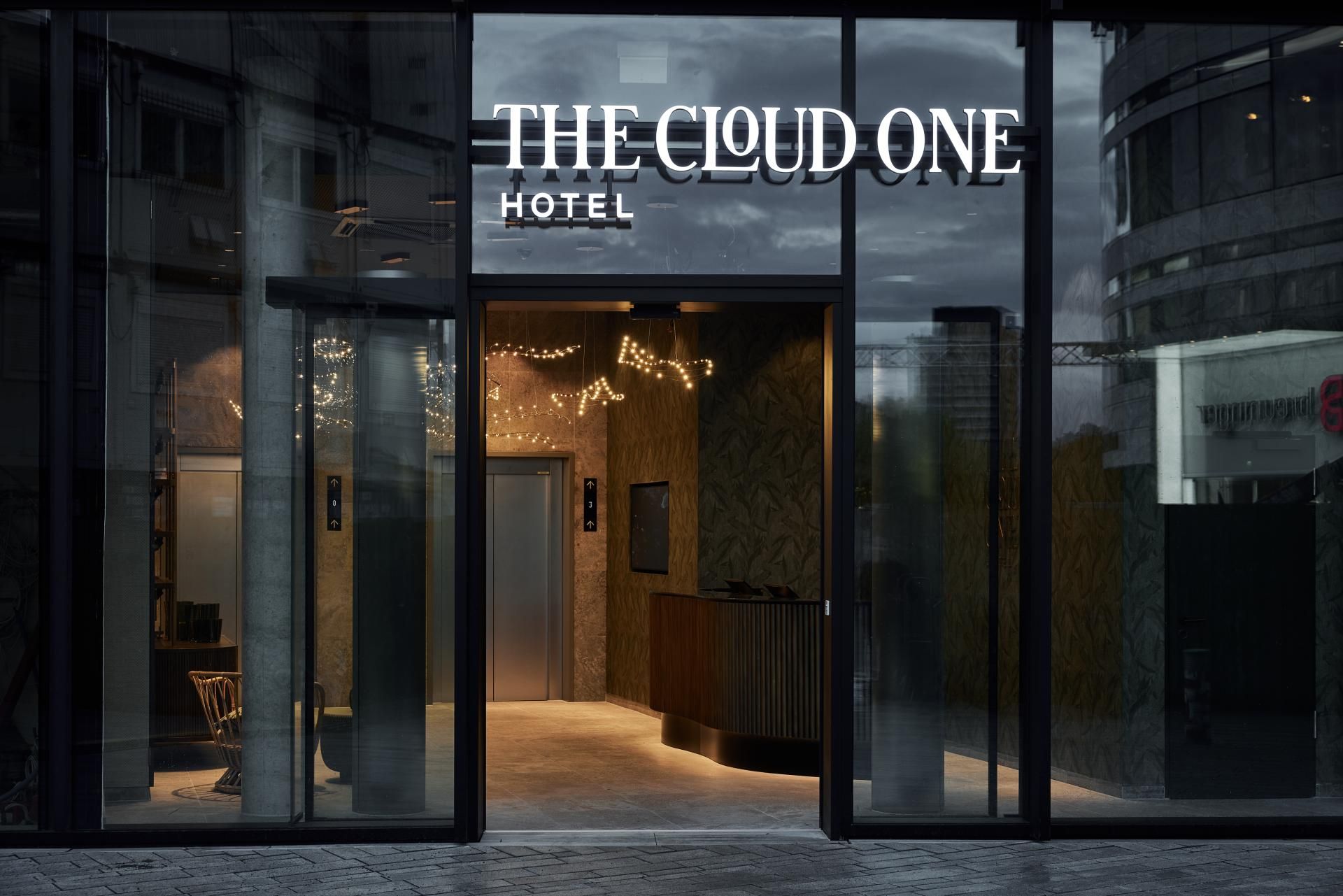 Moderne Hoteleingang mit gläserner Fassade, beleuchtetem Empfangsbereich und stilvollem Interieur. Logo „THE CLOUD ONE HOTEL“ prominent über dem Eingang angeleuchtet. Einladende Atmosphäre für Geschäfts- und Urlaubsreisende.
