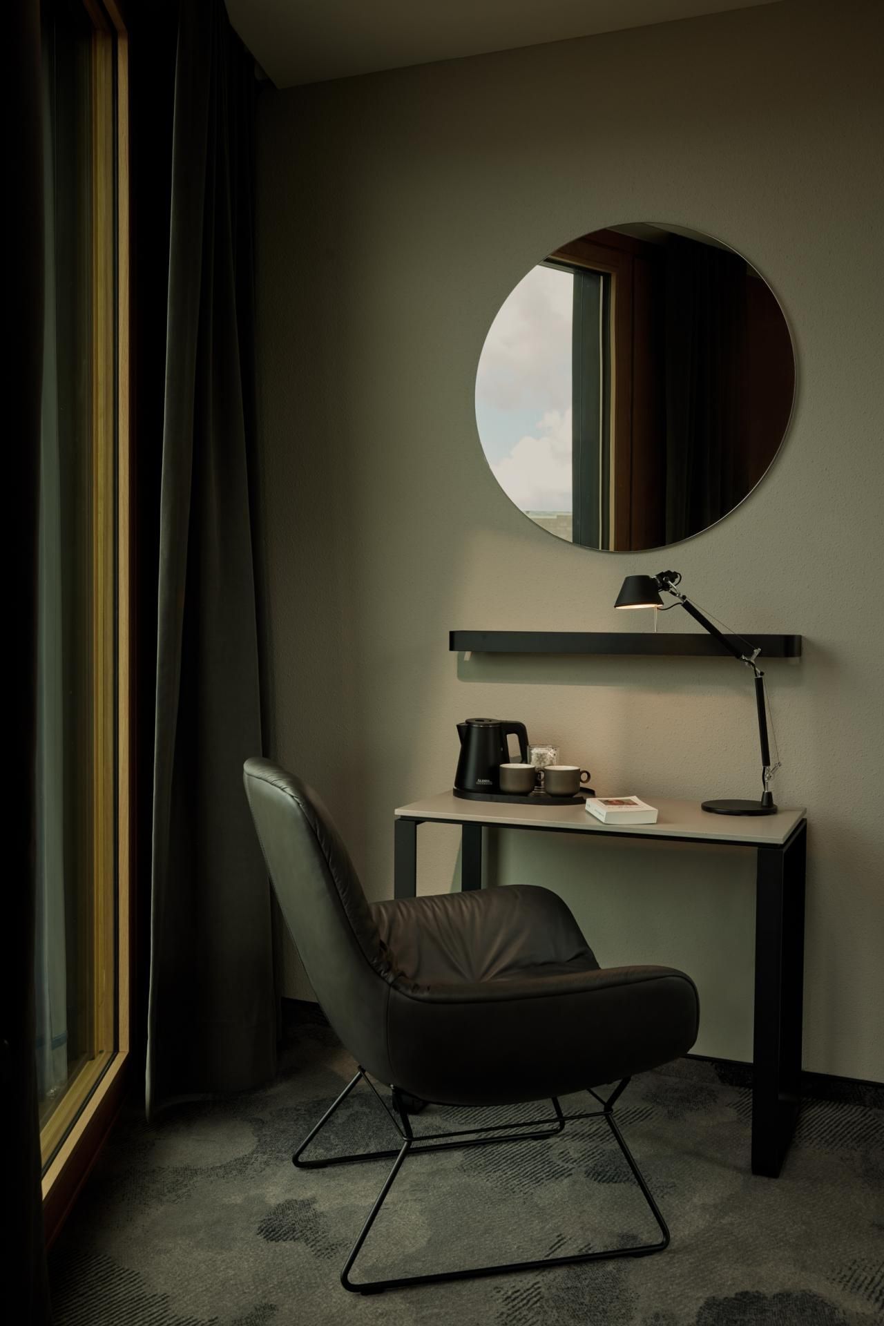 Moderner Arbeitsplatz im Hotelzimmer mit komfortablem Sessel, Schreibtisch, Leselampe, Teekocher und Tassen, dazu ein rundes Fenster für Tageslicht und Ausblick. Ruhige, einladende Atmosphäre für konzentriertes Arbeiten oder entspanntes Lesen.