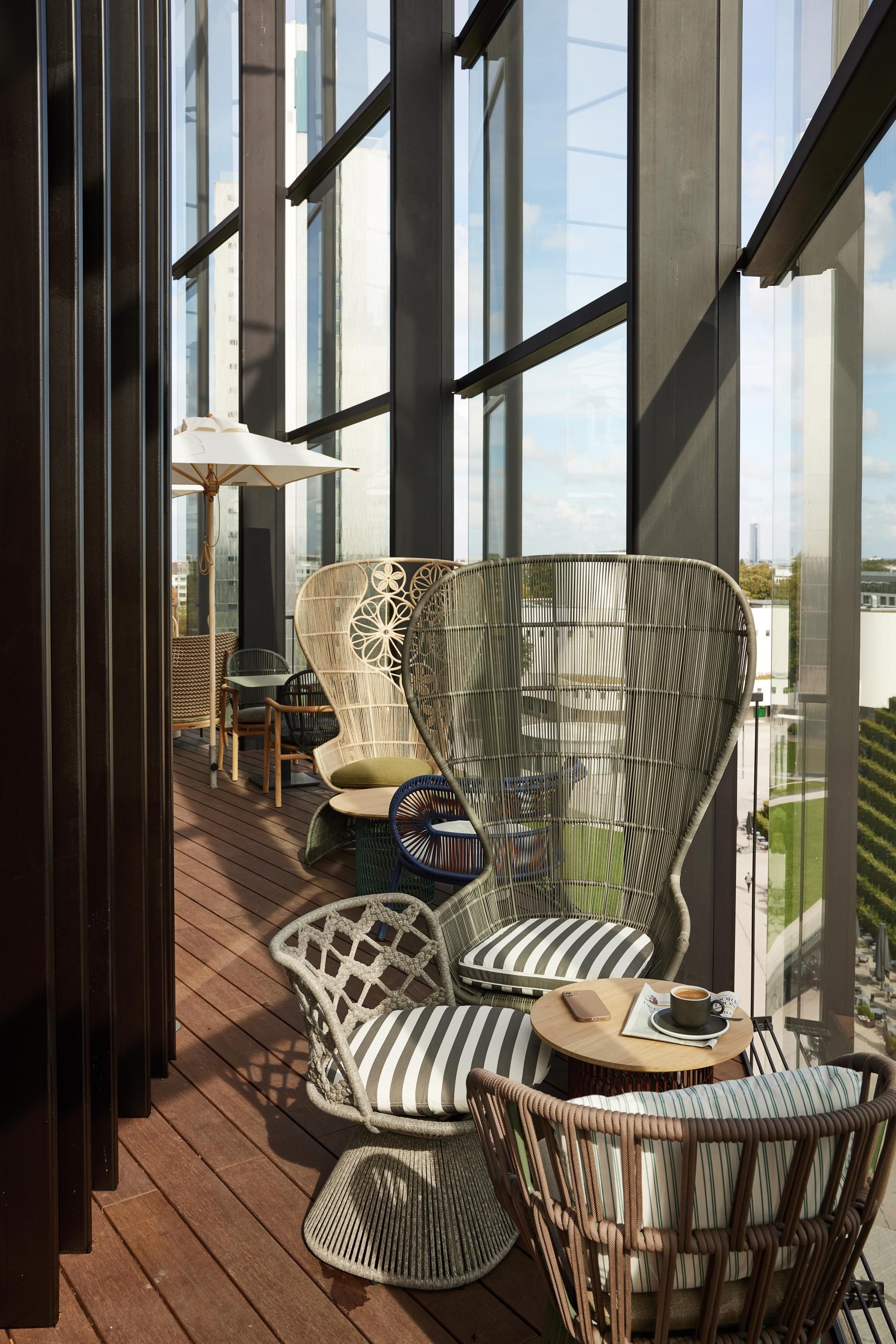 Balkon mit modernen, geflochtenen Sitzmöbeln und Kissen, Holzboden und großer Glasfront mit Blick auf die Stadt, gemütliche Atmosphäre für entspannte Momente.