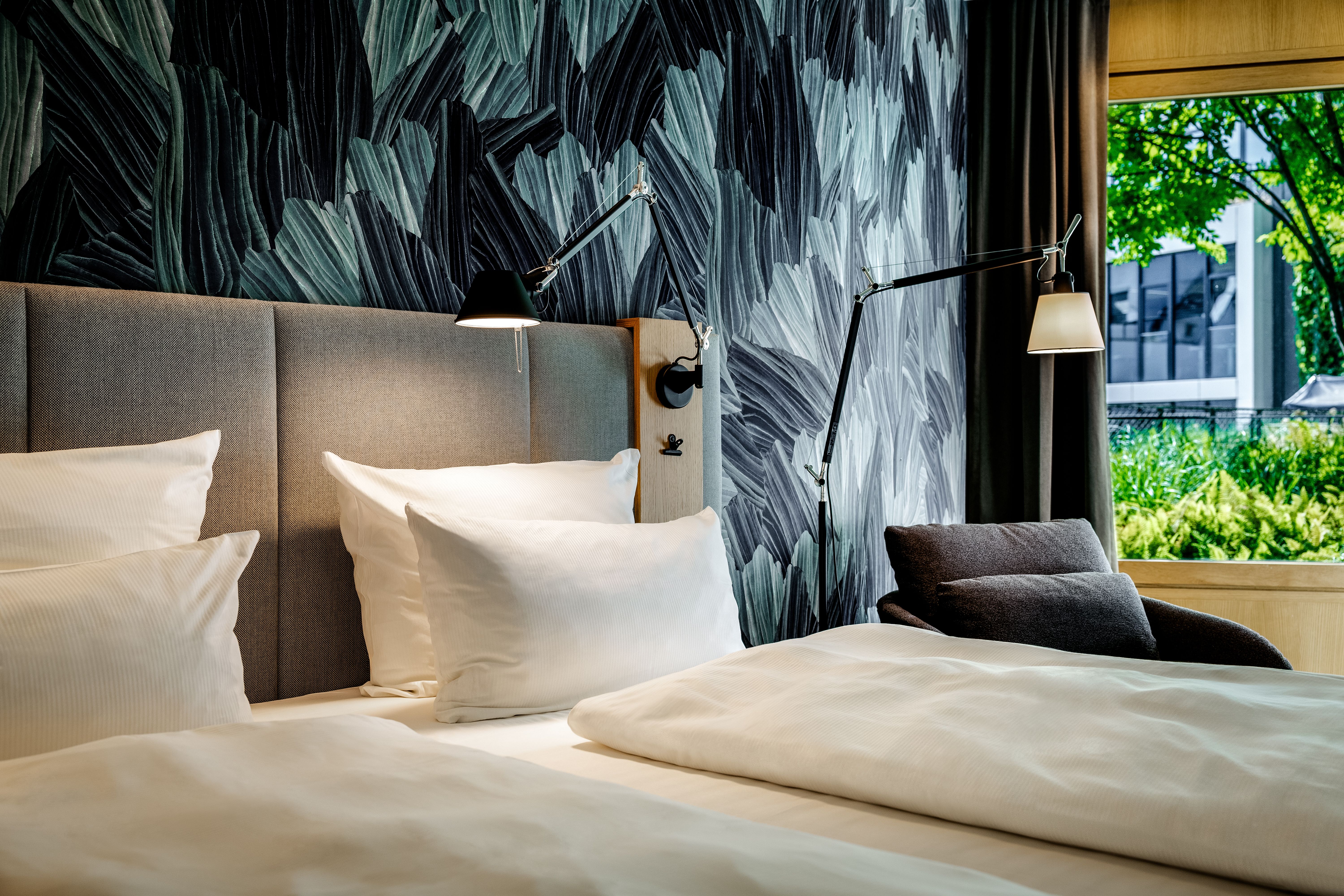 Gemütliches Doppelbett mit weißen Bettwäsche und Kopfkissen, moderner Wandbeleuchtung, stilvoll gemusterte Wand und großes Fenster mit Blick ins Grüne für entspannte Atmosphäre.