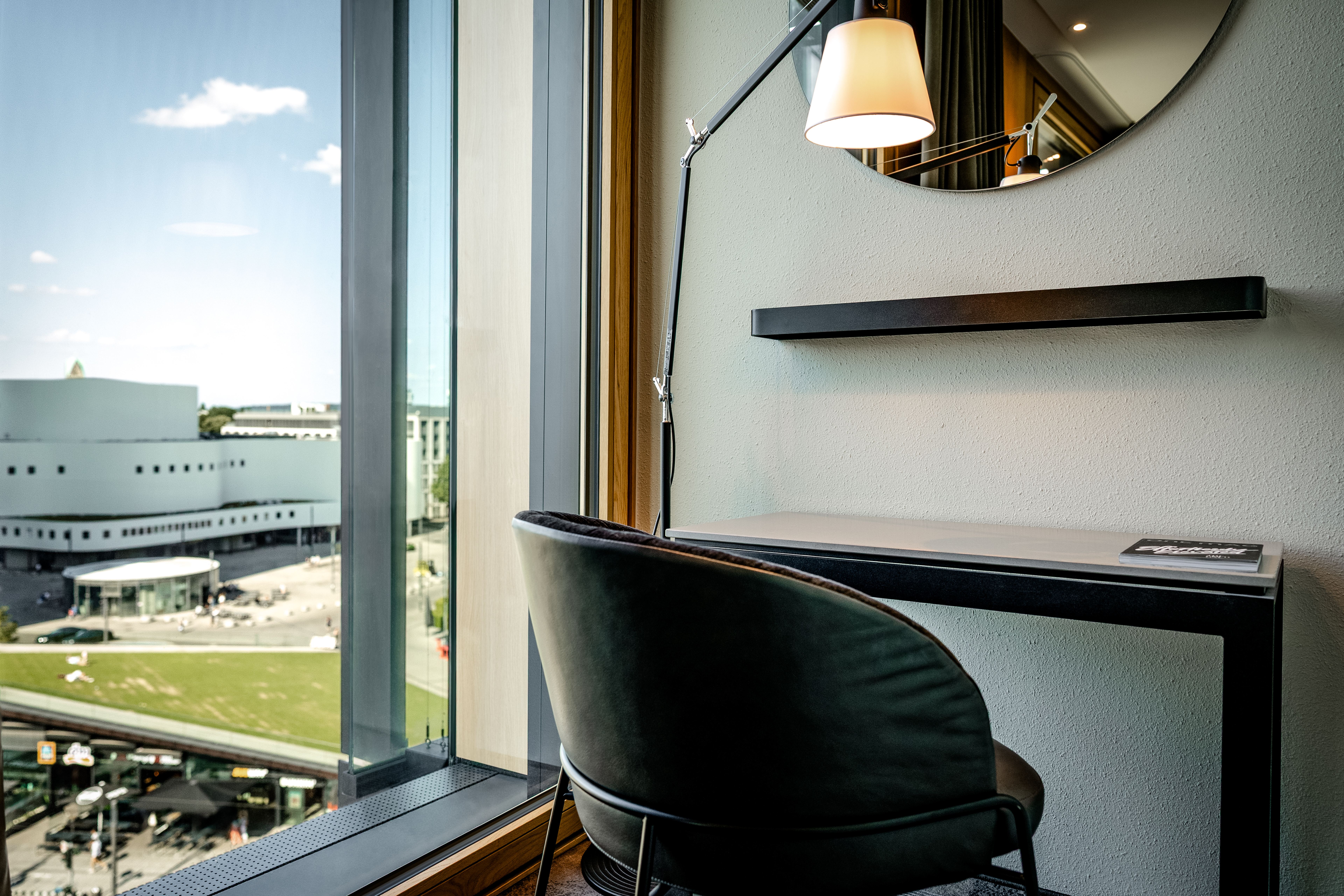 Moderner Arbeitsplatz am Fenster mit Blick auf die Stadt, komfortabler Stuhl, schlichte Arbeitsfläche, runde Wandspiegel und warme Beleuchtung schaffen eine ruhige, inspirierende Atmosphäre. Moderner Arbeitsplatz am Fenster mit Blick auf die Stadt, komfortabler Stuhl, schlichte Arbeitsfläche, runde Wandspiegel und warme Beleuchtung schaffen eine ruhige, inspirierende Atmosphäre.