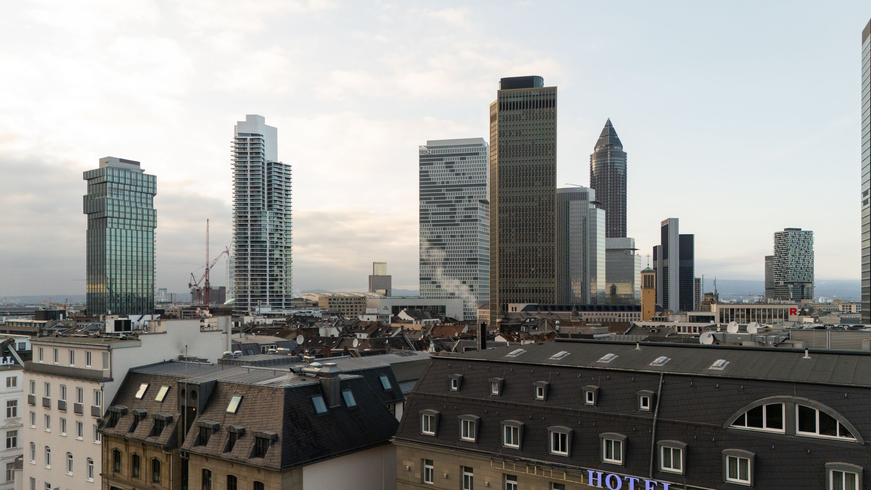 Hotel Frankfurt Metropolitan The Cloud One Blick auf die Skyline einer modernen Großstadt mit hohen Bürogebäuden und älteren Wohnhäusern im Vordergrund bei bewölktem Himmel.