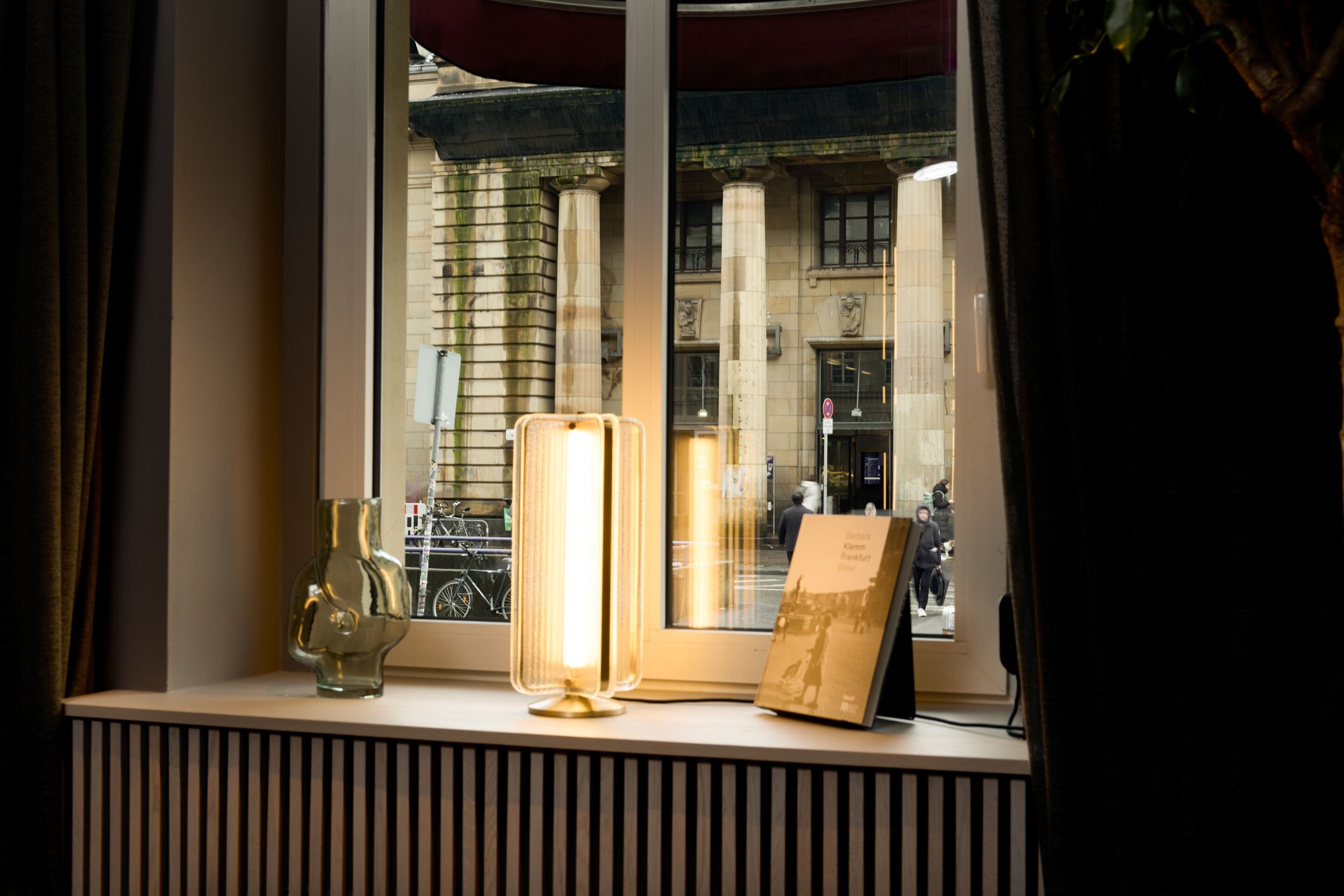 Designhotel Frankfurt Metropolitan The Cloud One Blick aus einem Zimmerfenster auf historische Säulen eines Gebäudes. Auf der Fensterbank steht eine Tischlampe, daneben eine Glasvase und ein aufgestelltes Buch.