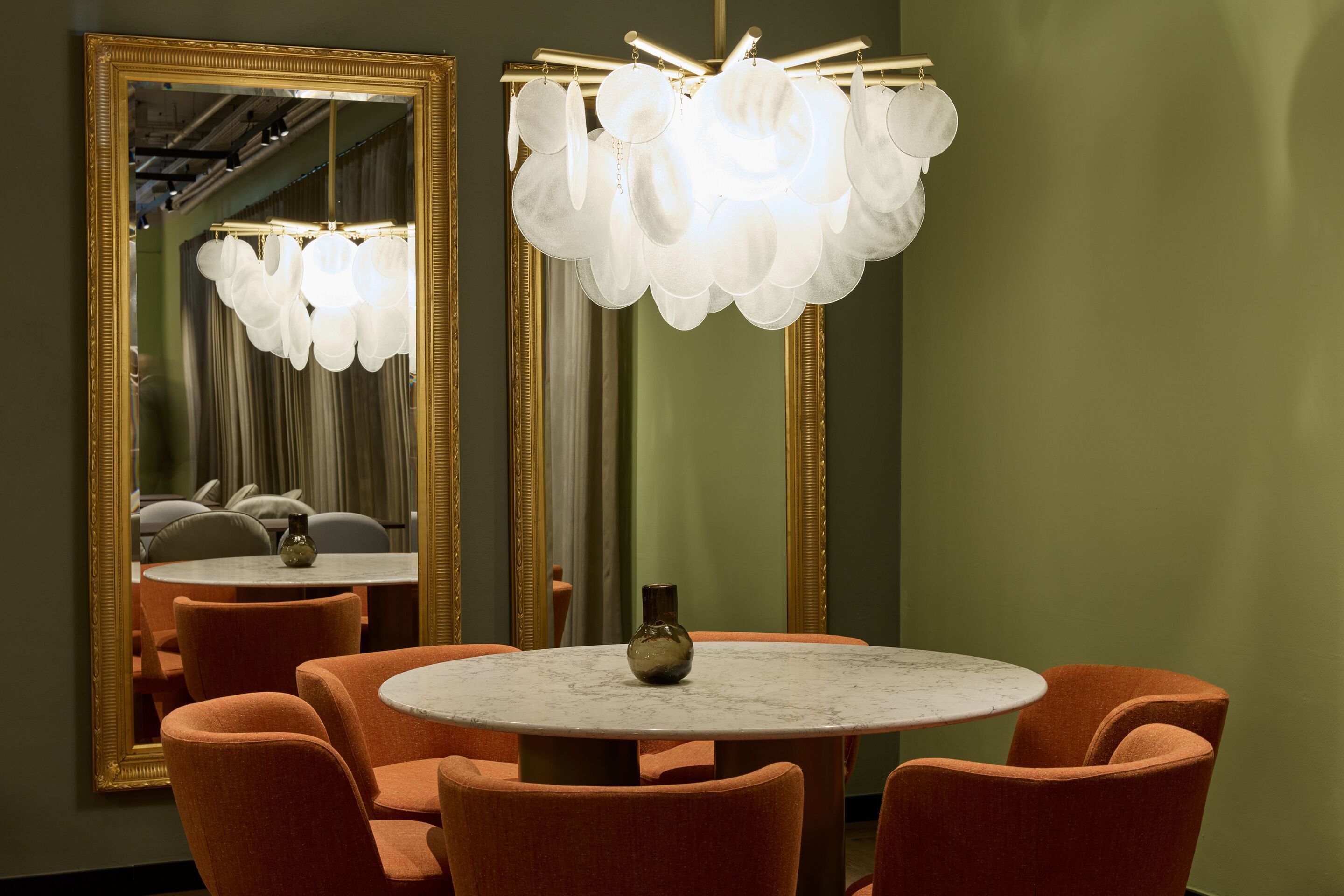 Hotel Frankfurt-Metropolitan The Cloud One Essbereich mit rundem Marmortisch, vier orangeroten Stühlen und einem markanten, runden Deckenleuchter aus weißen Scheiben vor einer olivgrünen Wand und großem Wandspiegel.