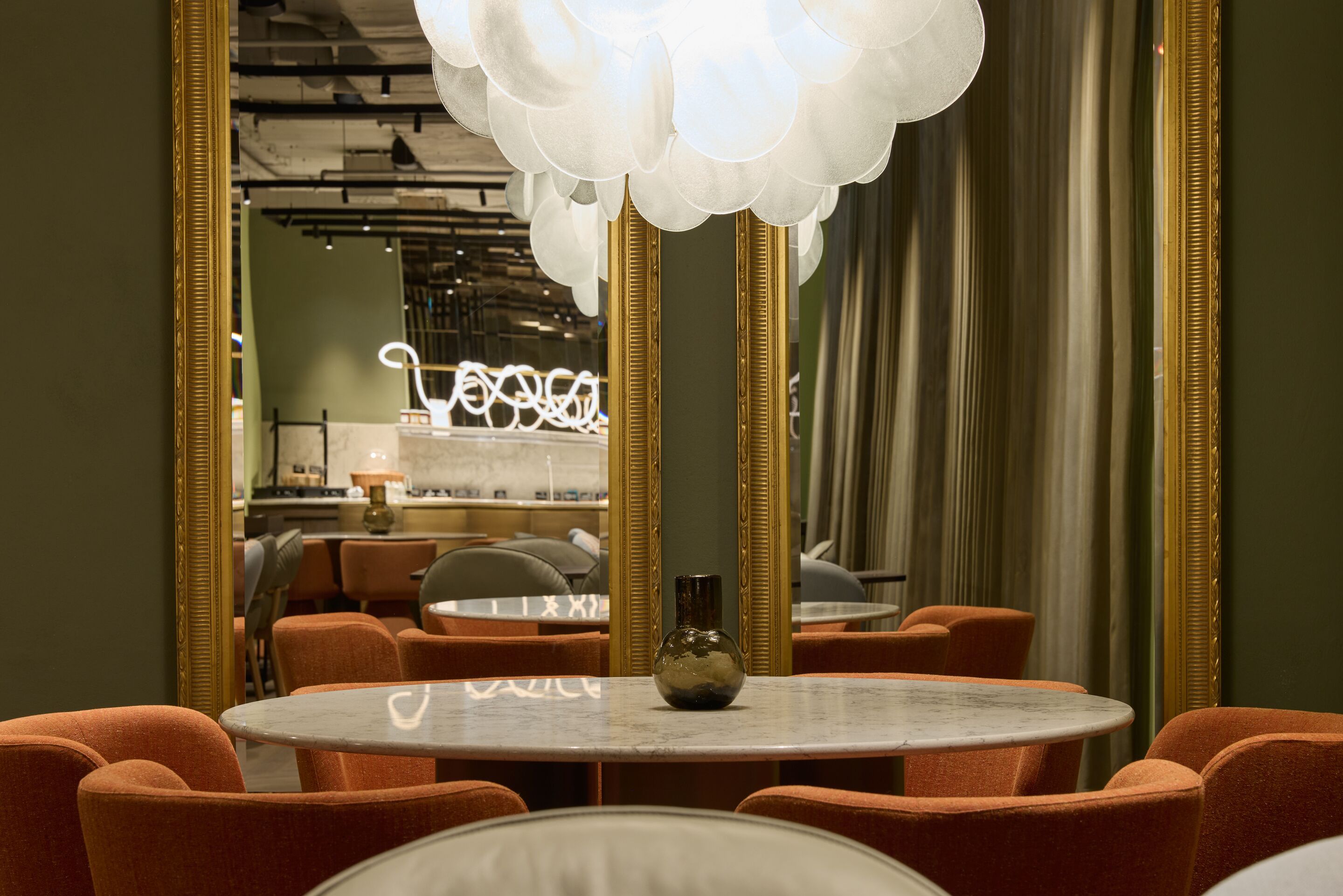 Hotel Frankfurt-Metropolitan The Cloud One Sitzbereich mit rundem Marmor-Tisch, orangen Polstersesseln und großzügigem Spiegel, der den Raum optisch erweitert. Sanfte Beleuchtung durch eine moderne, wolkenartige Hängeleuchte.
