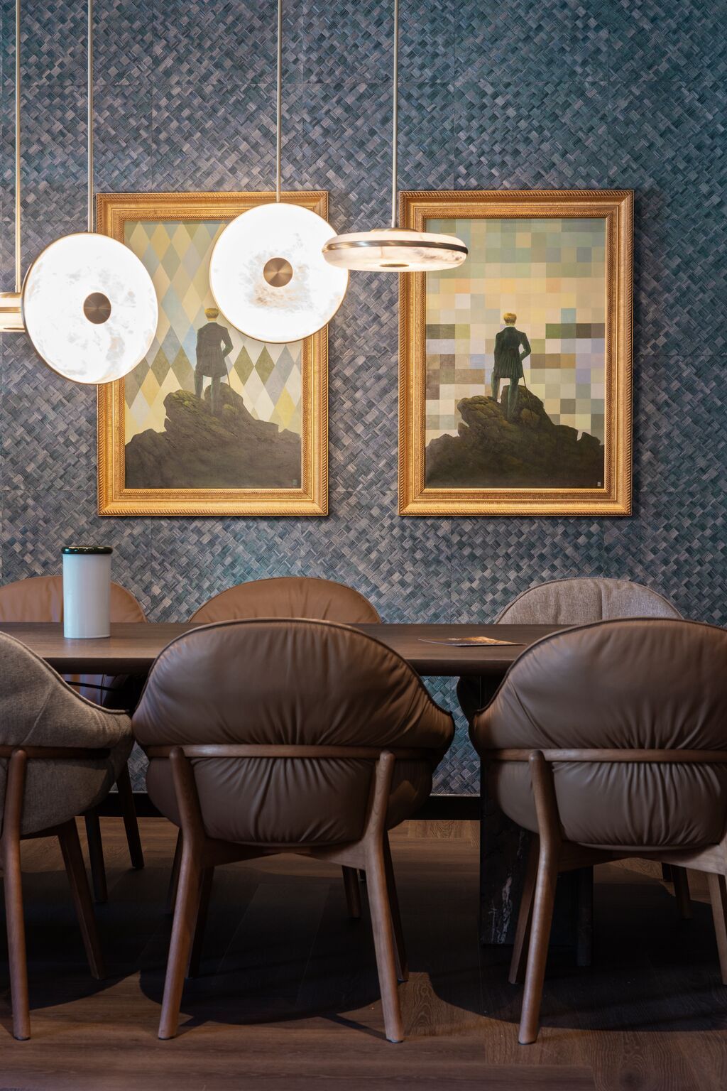 Designhotel Frankfurt-Metropolitan The Cloud One Tische und Stühle mit braunen Lederstühlen, einem Holztisch und stilvollen Pendelleuchten, im Hintergrund zwei gerahmte Kunstwerke mit stilisierten Figuren vor geometrischen Hintergründen.