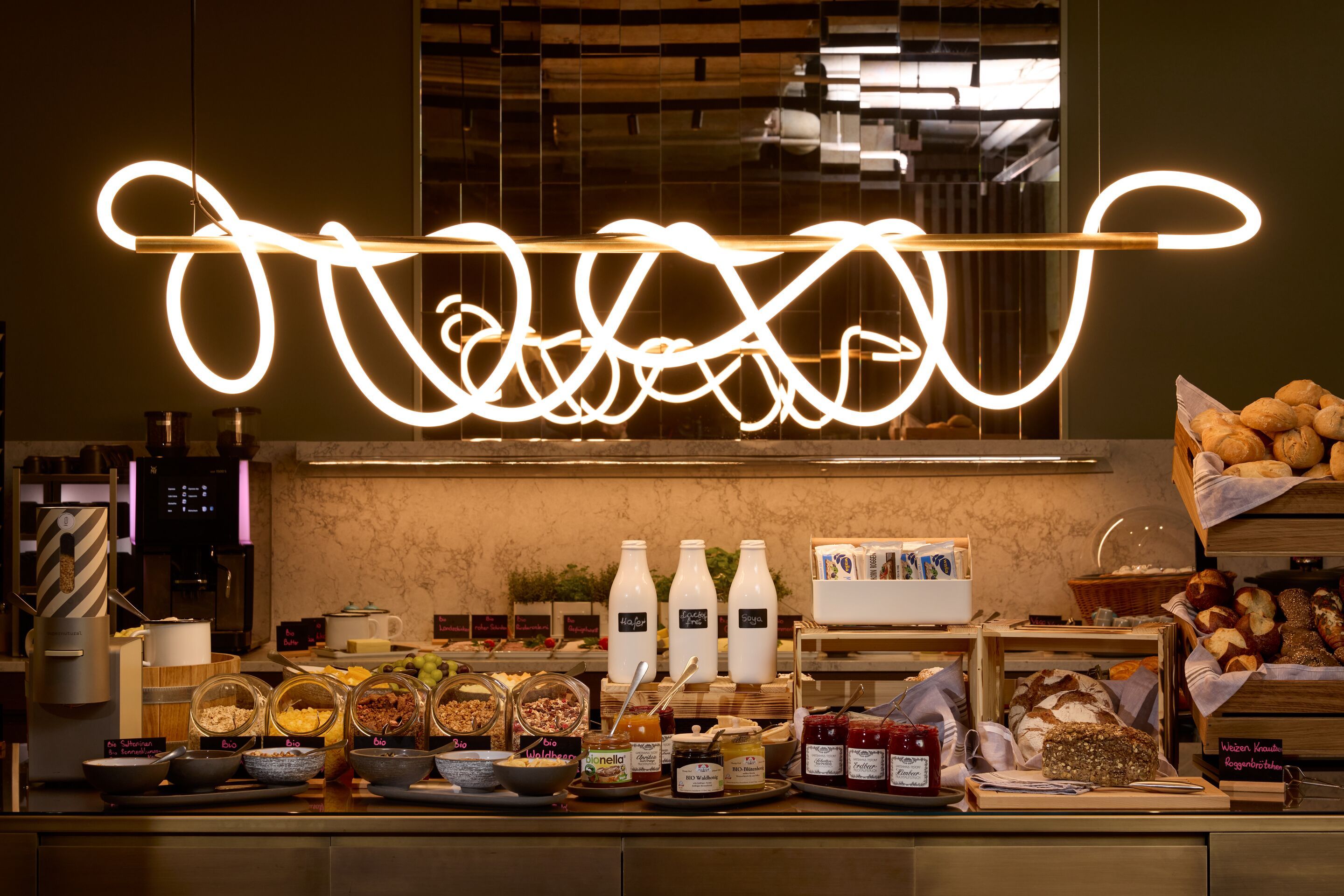 Hotel Frankfurt Metropolitan The Cloud One hell beleuchtetes Frühstücksbuffet mit verschiedenen Müslisorten, frischem Brot, Aufstrichen und Milchflaschen in einem modernen, einladenden Ambiente.