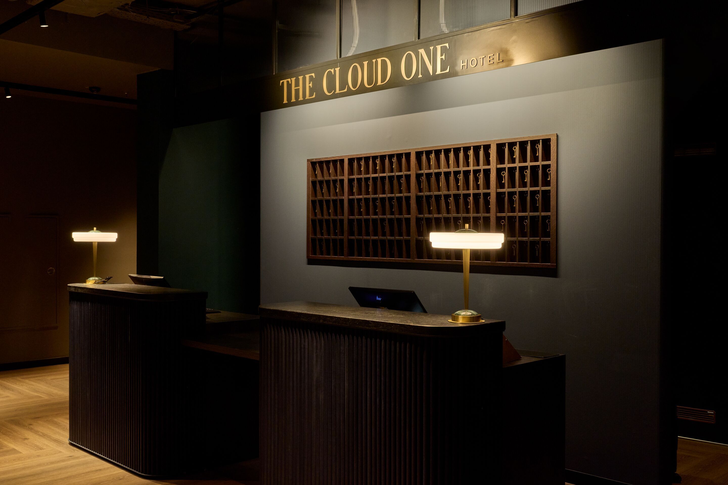Hotel Frankfurt Metropolitan The Cloud One Rezeption eines mit dunklem Holz, warmem Licht und einer Wand mit Schlüsselfächern, die für ein einladendes Ambiente und professionellen Service steht.