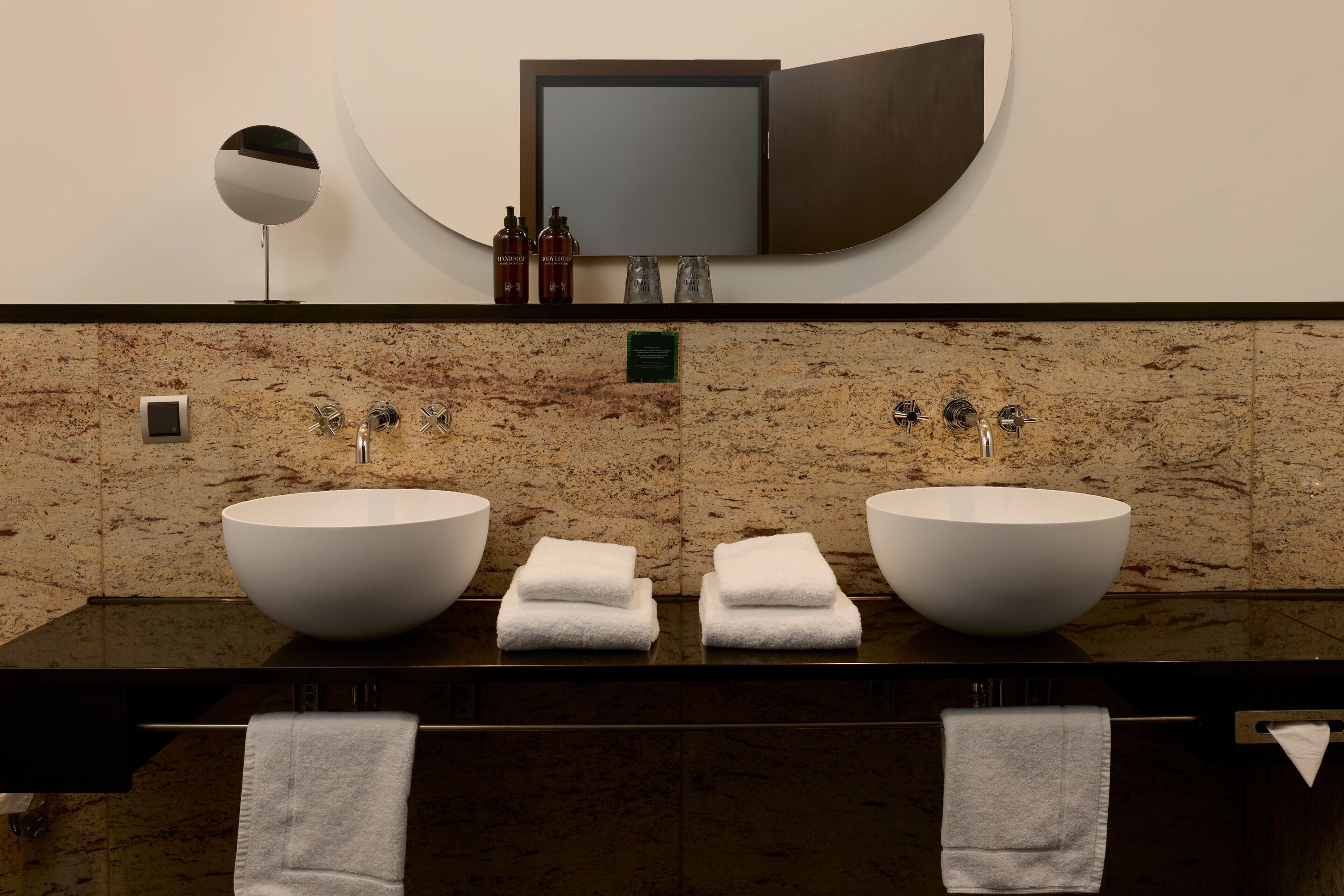 Designhotel Frankfurt Metropolitan The Cloud One Badezimmer mit zwei runden freistehenden Waschbecken, weißen Handtüchern und einer großen, ovalen Wandspiegel. 