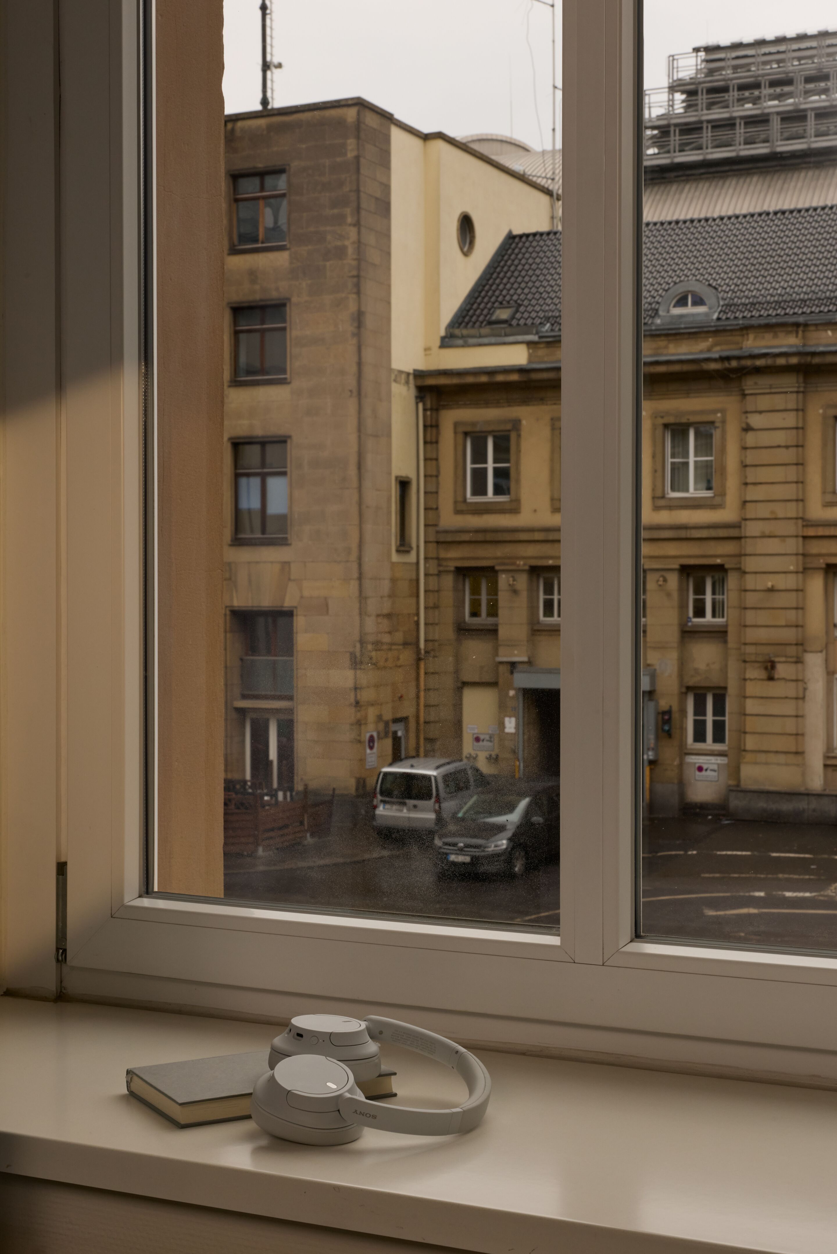Hotel Frankfurt Metropolitan The Cloud One Blick aus einem Zimmerfenster auf gegenüberliegendes Gebäude und geparkte Autos. Auf der Fensterbank liegen ein Buch und ein weißer Kopfhörer, die eine entspanntes Ambiente zum Lesen oder Musik hören vermitteln.